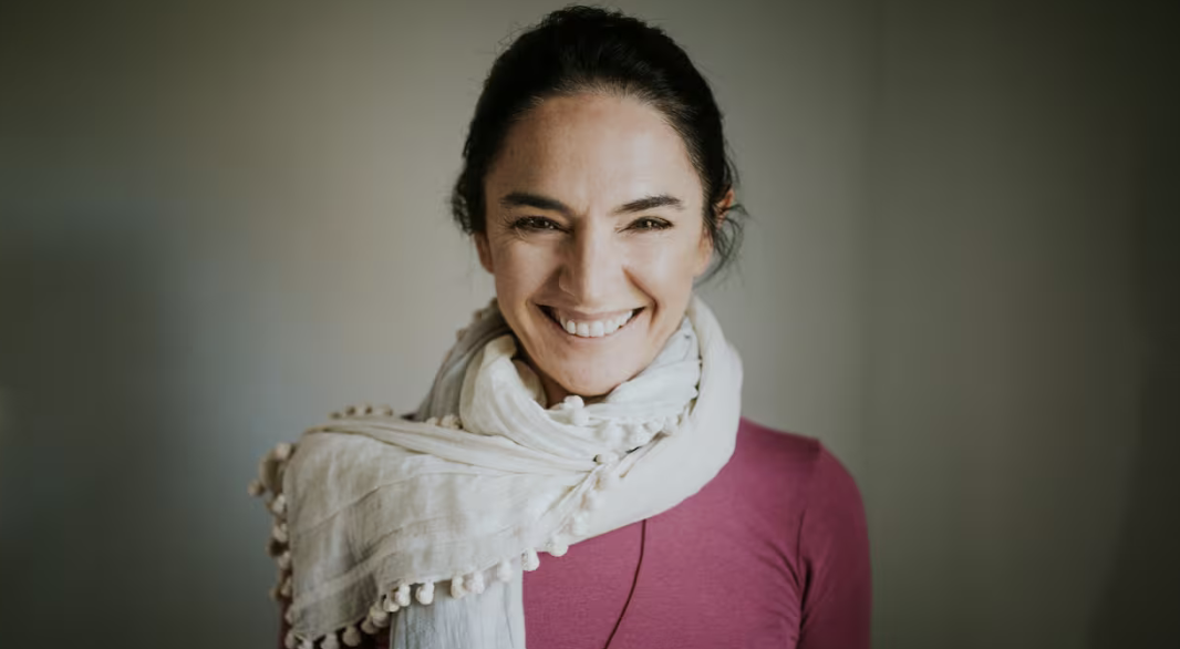 A woman smiling, wearing a pink top and a white scarf in front of a plain background.