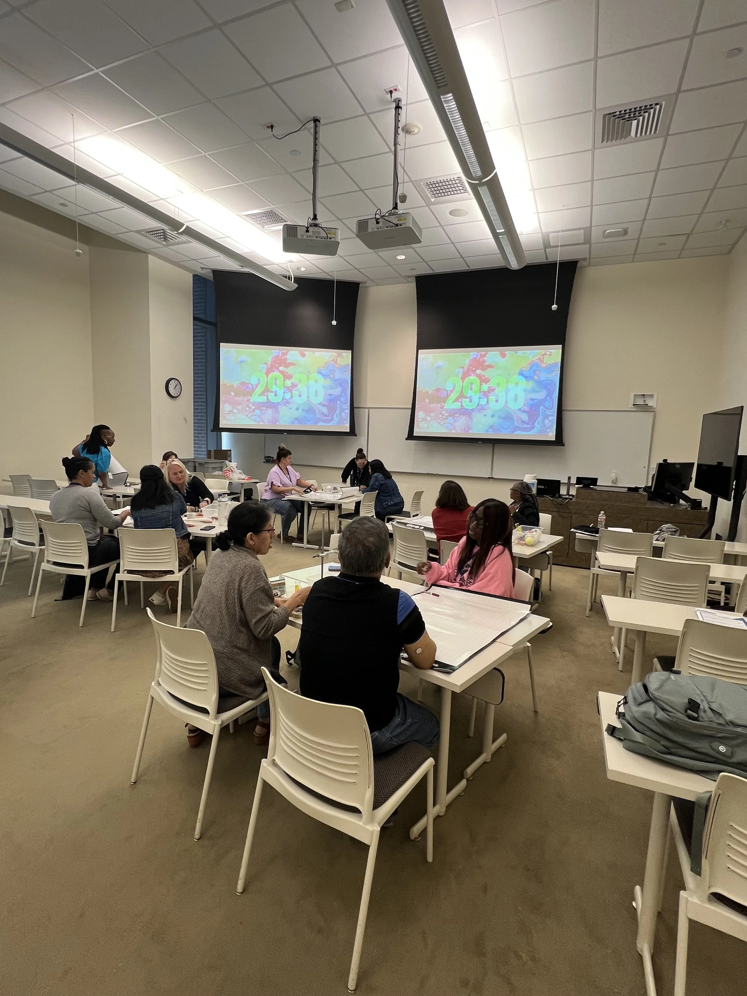 A classroom or conference room with people sitting at tables, two large projection screens displaying a digital timer, some people wearing face masks, and computers on the desks.