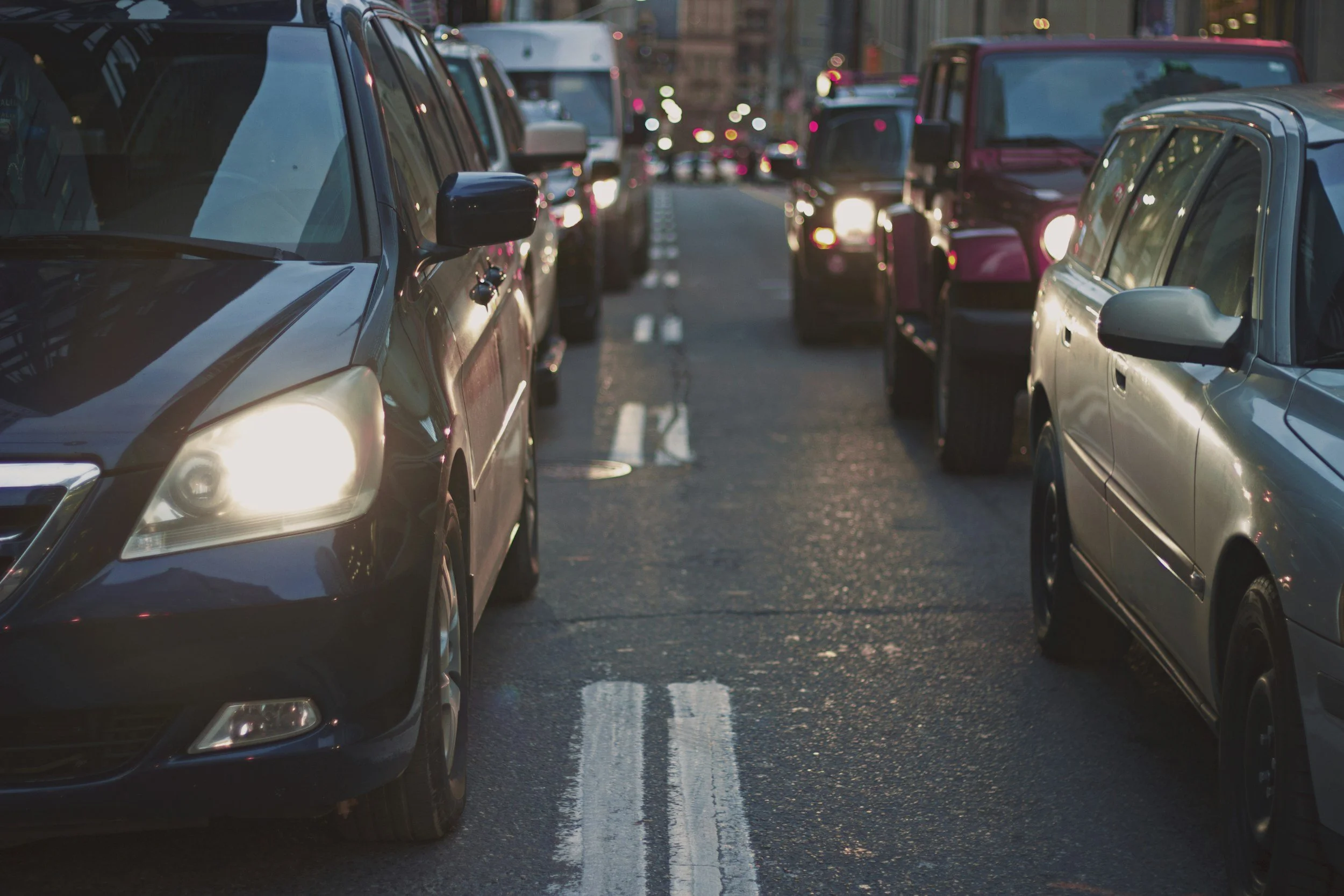 Multiple parked cars along a city street at dusk with reflections on the wet pavement.
