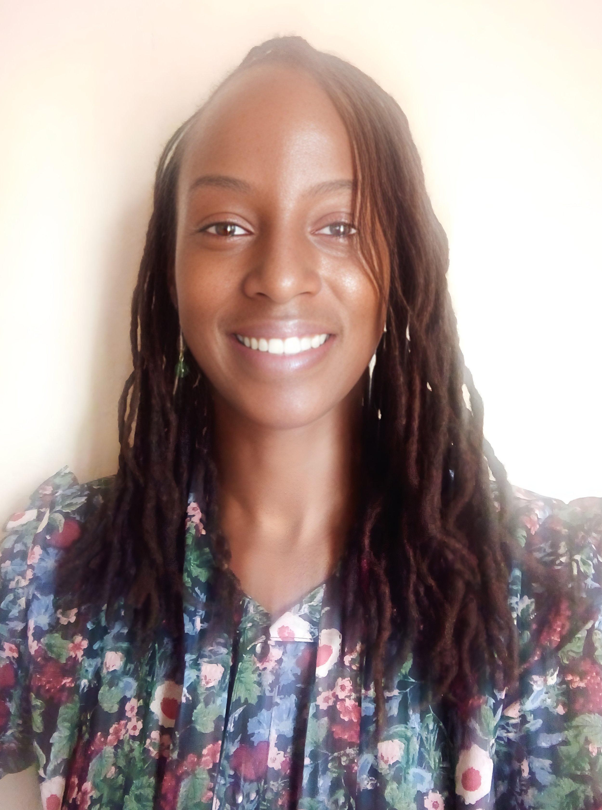 A woman with medium skin tone and long, dark, curly dreadlocks, smiling, wearing a floral blouse, standing against a beige wall.