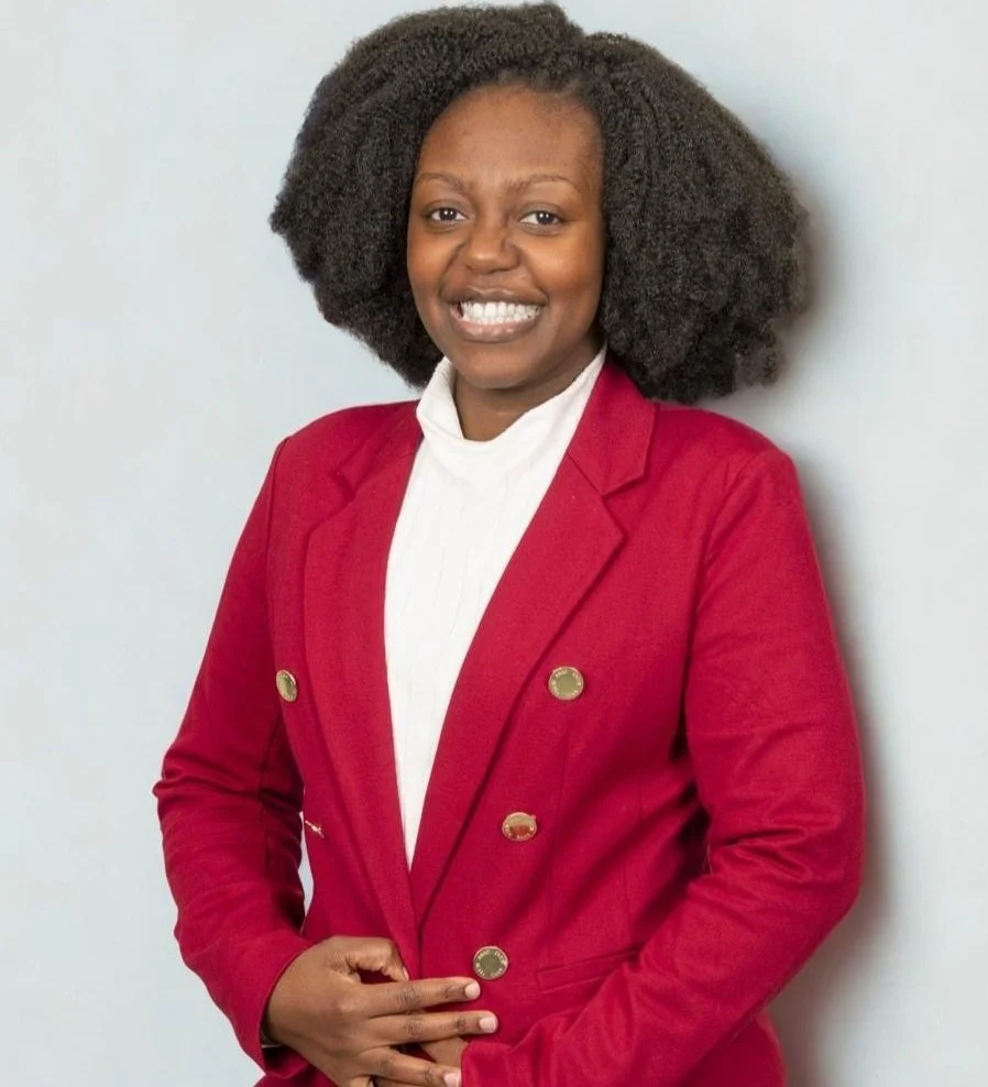 woman with natural curly hair wearing a red blazer and white blouse, standing against a neutral background, smiling at the camera