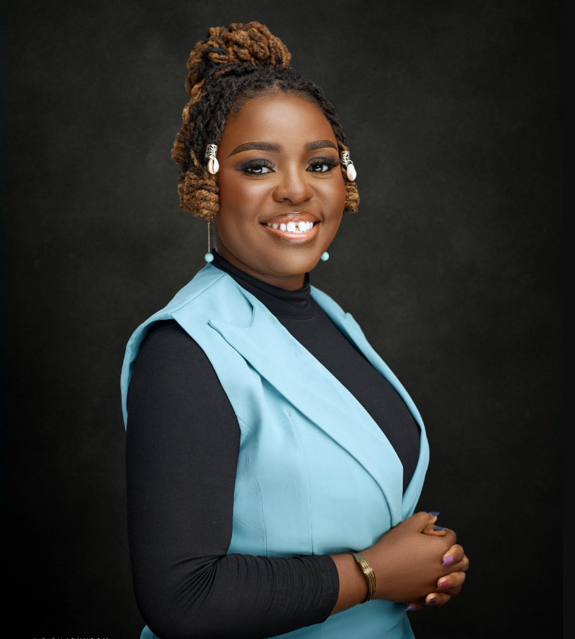 A smiling Black woman with styled dreadlocks wearing a black top and a light blue vest against a dark background.