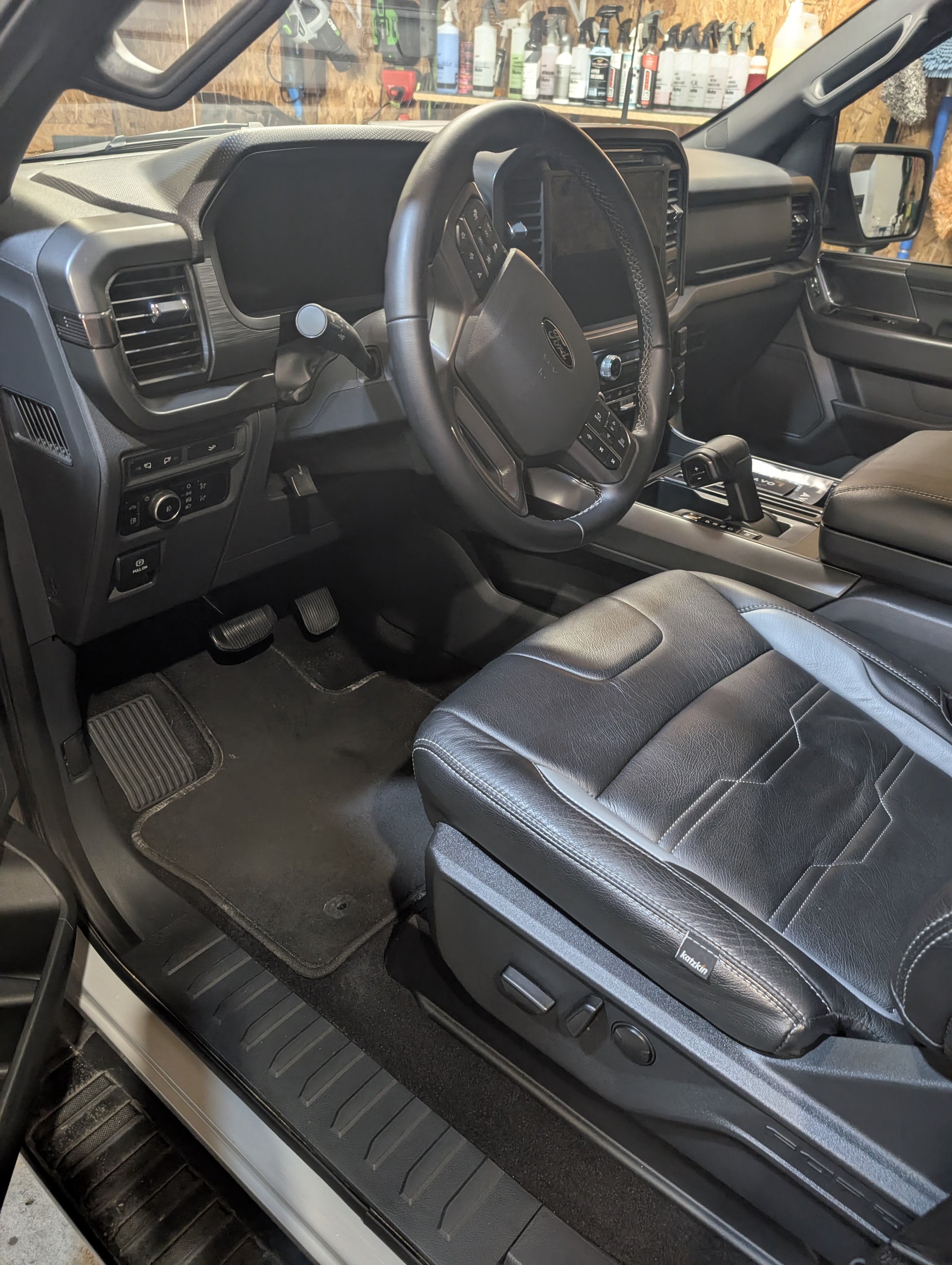 Interior of a black Ford truck showing the driver's seat, steering wheel, dashboard with touchscreen, and gear shift. The garage in the background has shelves with car care products.