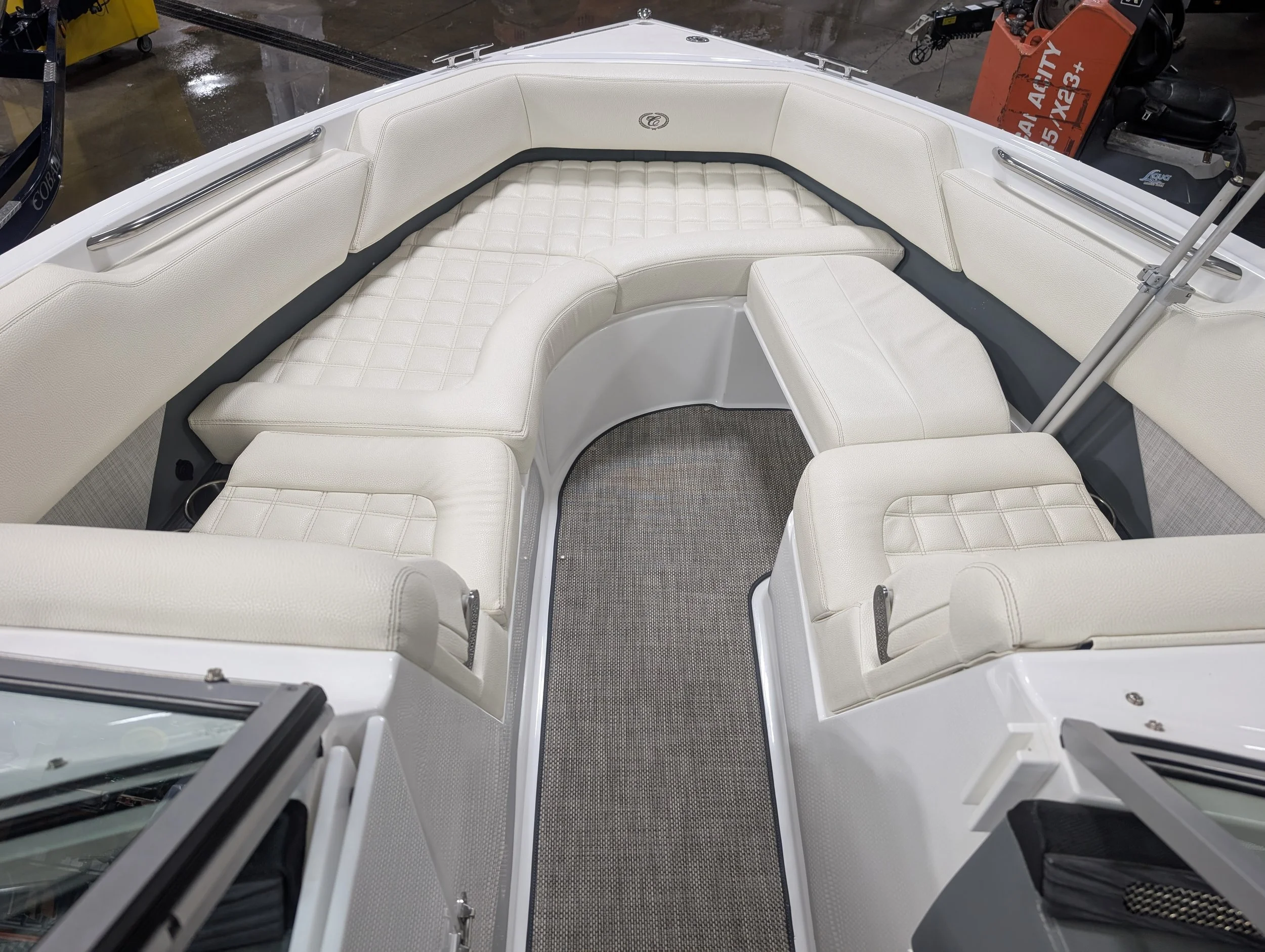 Interior view of a boat with white cushioned seating arranged in an L-shape, a small circular table surface, and a gray textured floor mat.