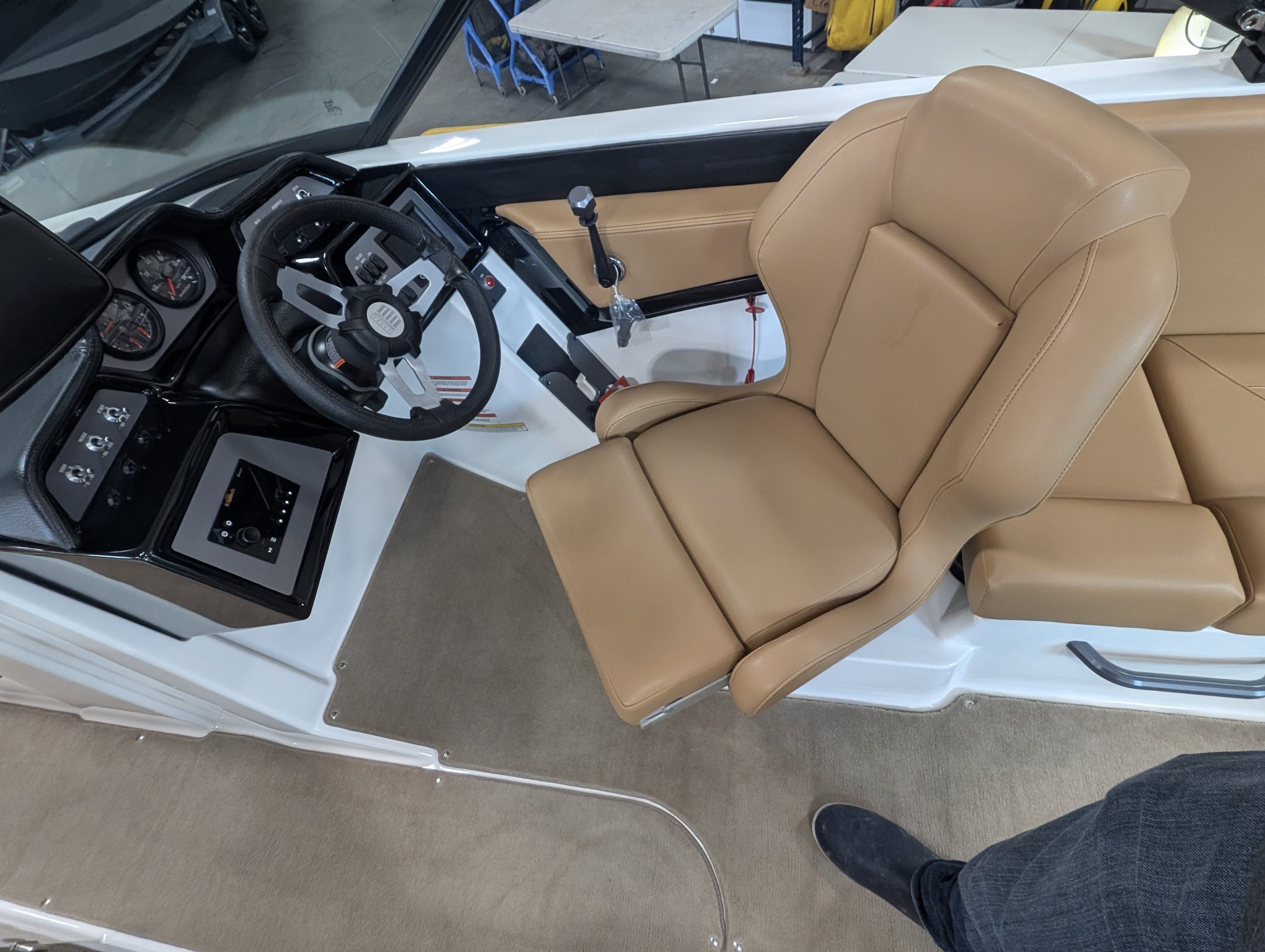 Inside view of a boat cockpit with a beige captain's chair, steering wheel, control panels, and a gear shift, with part of a person's foot visible.