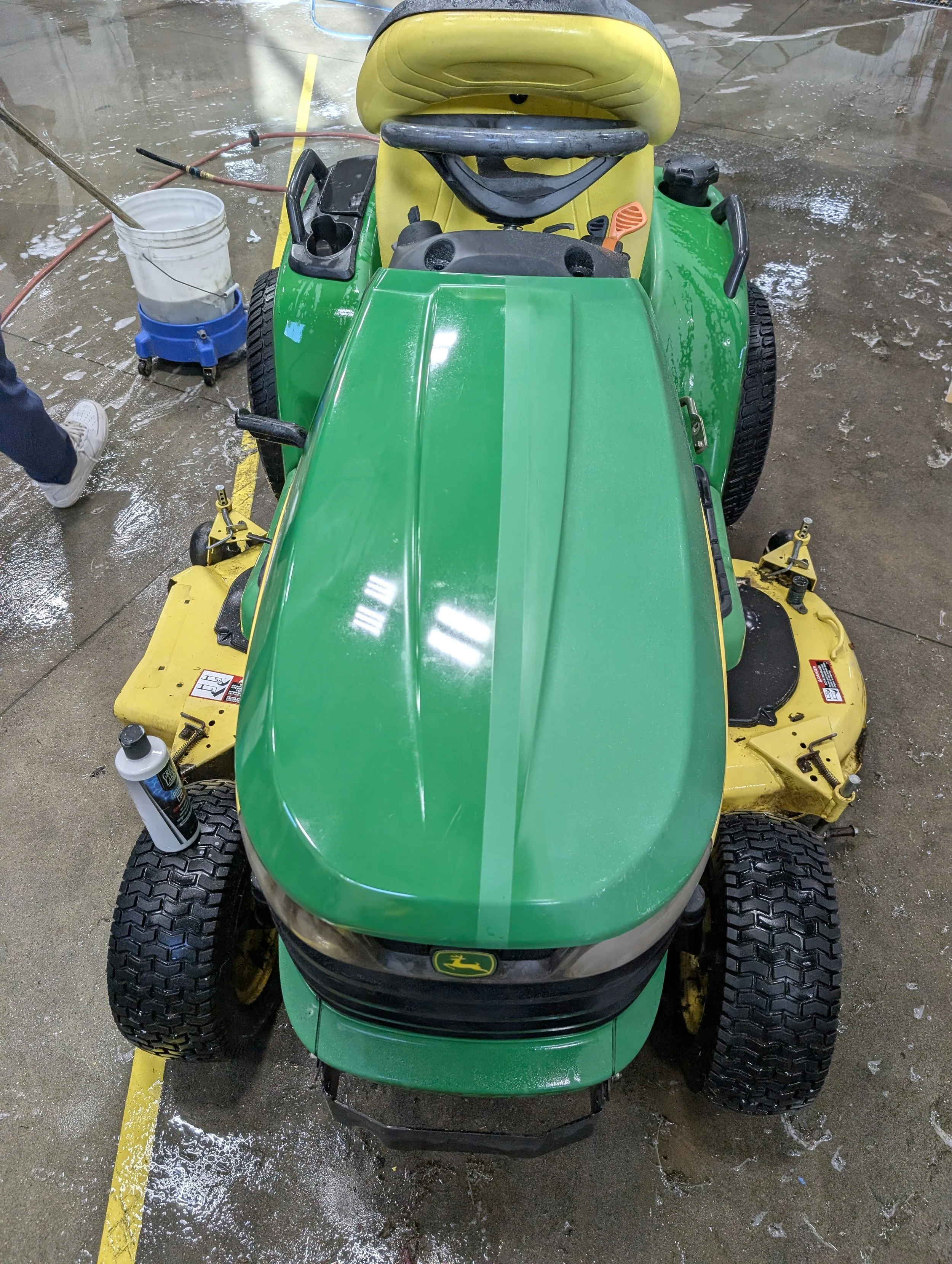 Green and yellow John Deere riding lawn mower inside a building, wet floor, cleaning supplies nearby.