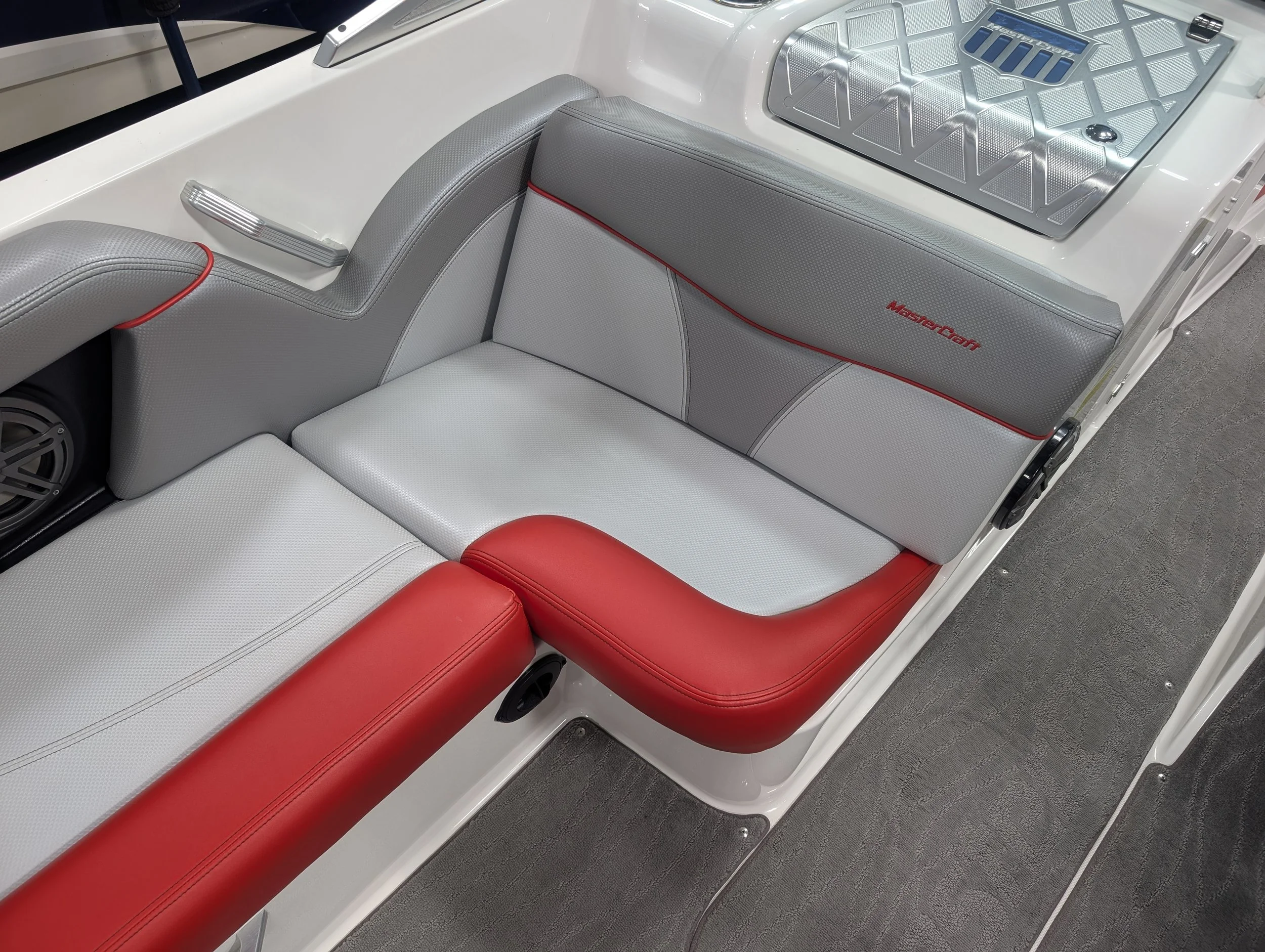 Close-up view of the interior of a small aircraft or boat with cushioned seats in gray, white, and red, with the words 'MasterCraft' embroidered on the gray cushion, a metal panel, and a speaker.