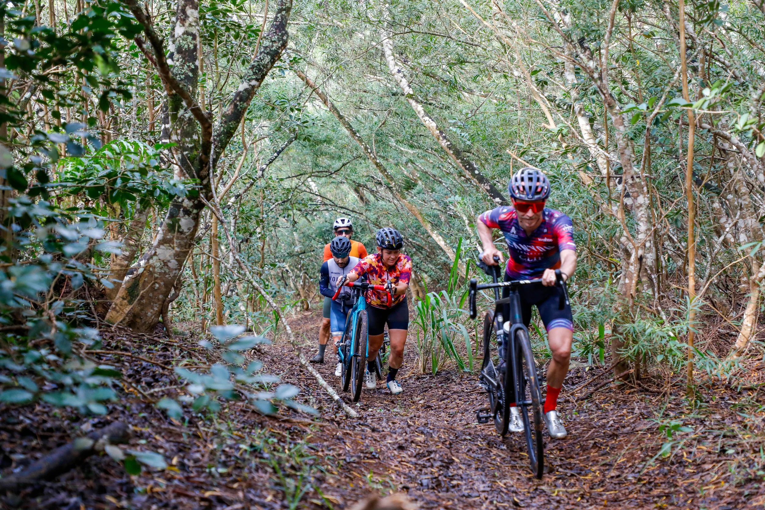 Four cyclists pushing their bikes uphill through a wooded trail, wearing cycling gear and helmets, surrounded by trees and foliage. Aloha Gravel. Hawaii Bike Race. Hawaii Gravel Bike