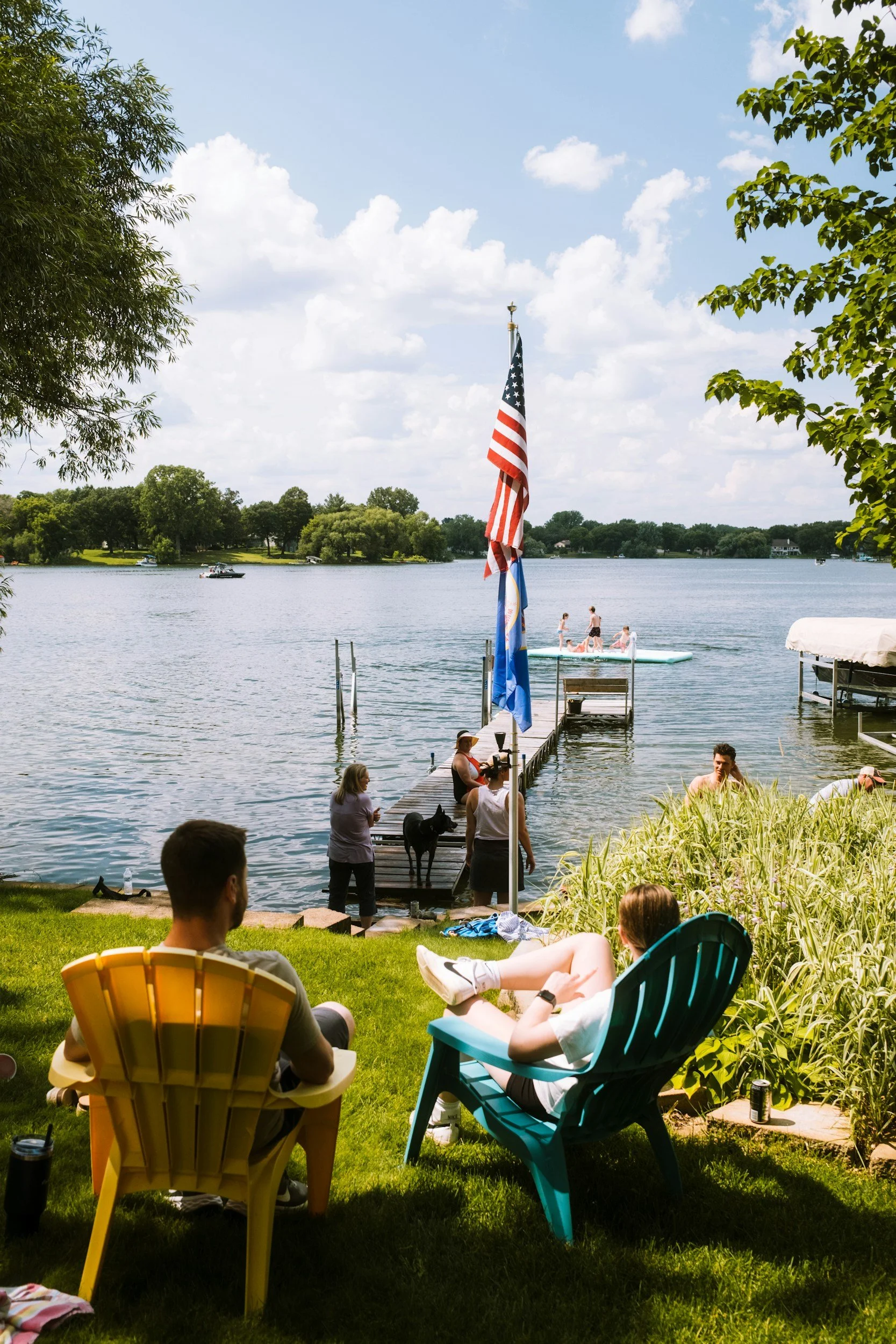 People relaxing by a lakeside on a sunny day, with two individuals sitting on lawn chairs, a dock extending into the water, an American flag and a blue flag on a pole, people swimming and boating in the lake, trees lining the shore, and a few clouds in the sky.