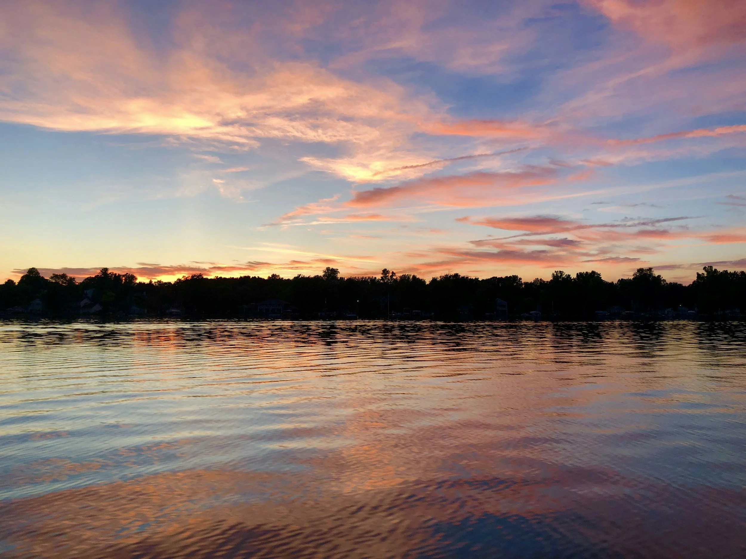 Sunset over a calm lake with trees along the shoreline, colorful clouds in the sky reflecting on the water.