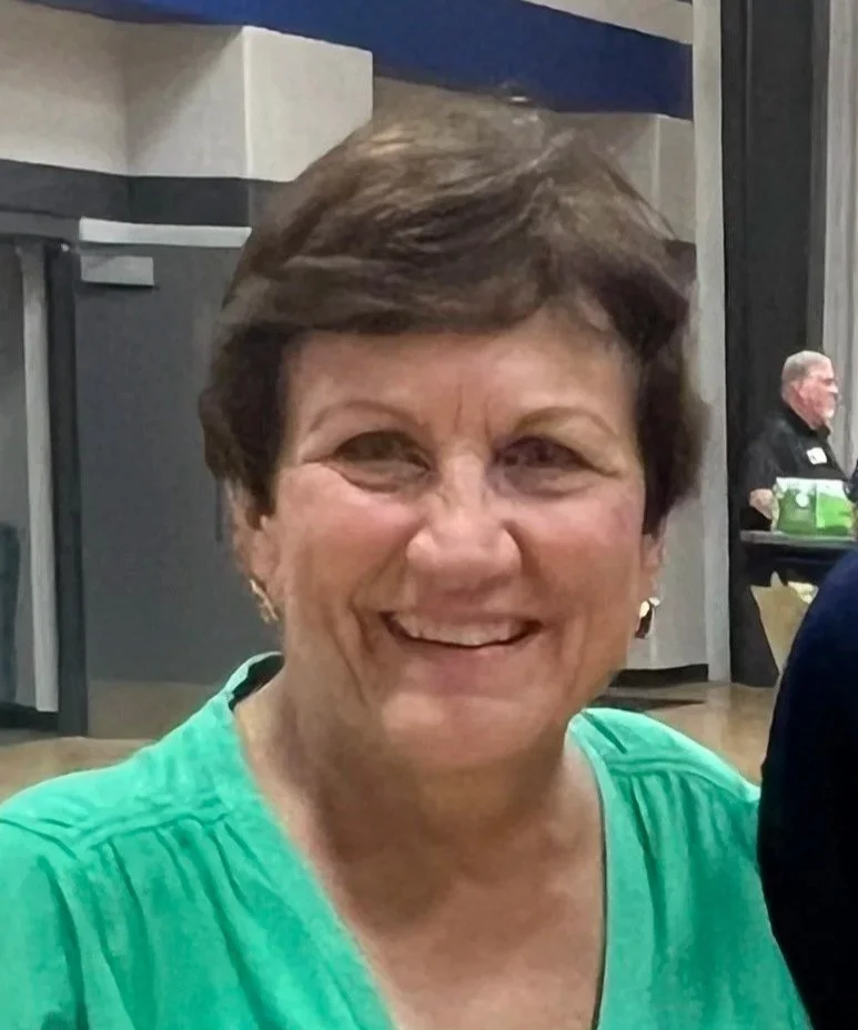 A smiling middle-aged woman with short brown hair wearing a green top, indoors in a modern setting, possibly a restaurant or event space.