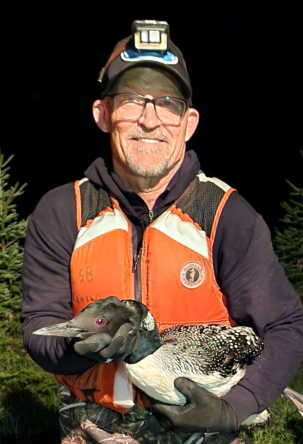 A man wearing a headlamp, glasses, and outdoor gear holding a wood duck, standing outdoors at night.