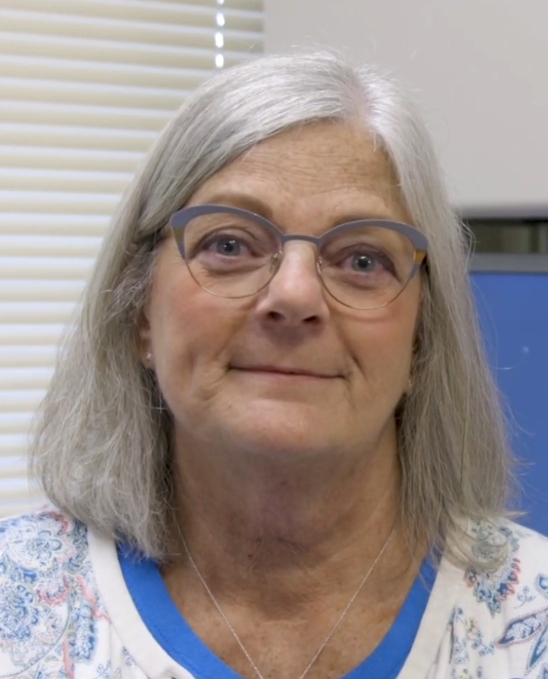 A middle-aged woman with gray hair, glasses, and a friendly smile, wearing a patterned shirt and a necklace, standing indoors in front of blinds and a blue screen or monitor.