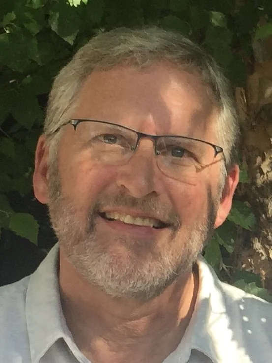 A middle-aged man with gray hair and a beard, wearing glasses and a light-colored shirt, smiling outdoors with green foliage in the background.