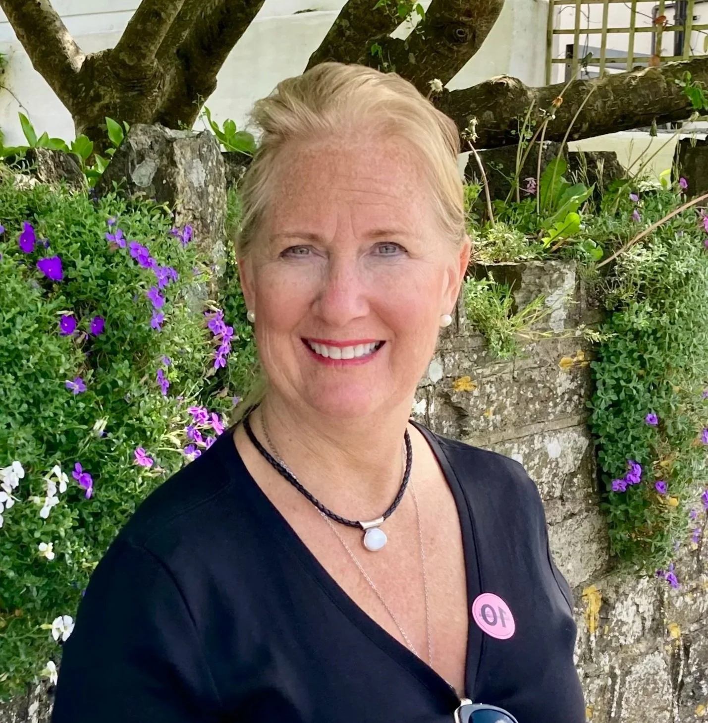 A smiling woman with blonde hair in loose waves, wearing pearls, a black top, and a pink '10' badge, standing in front of a stone wall adorned with purple and white flowers and green foliage.