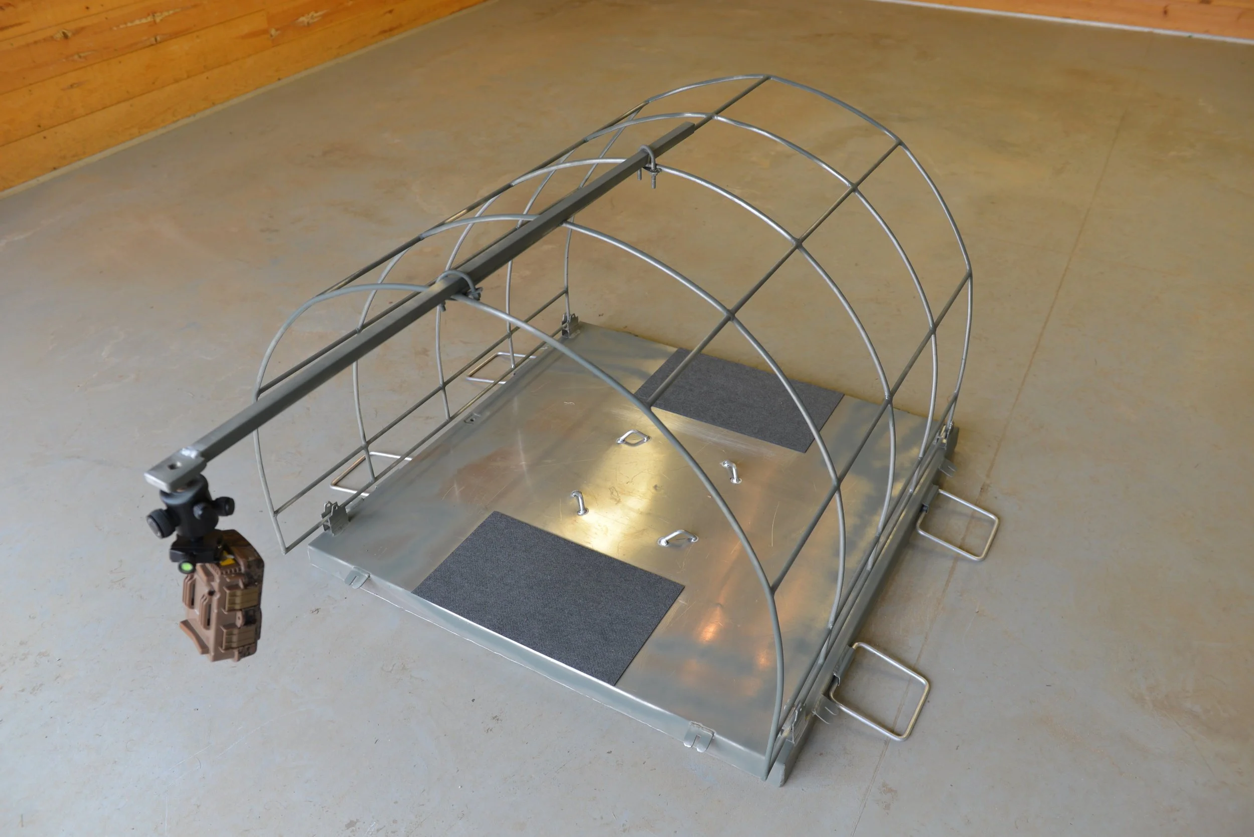 A metal dog crate with a sliding tray, two gray mats, and a camera mounted on a pole at the front.