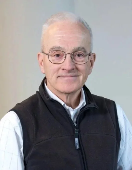 Older man with glasses, gray hair, wearing a white shirt and black vest, standing against a plain wall.