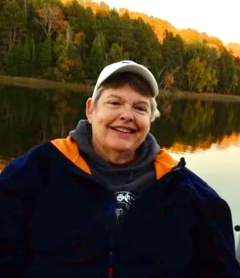 Person smiling outdoors near a lake with trees and fall foliage in the background.
