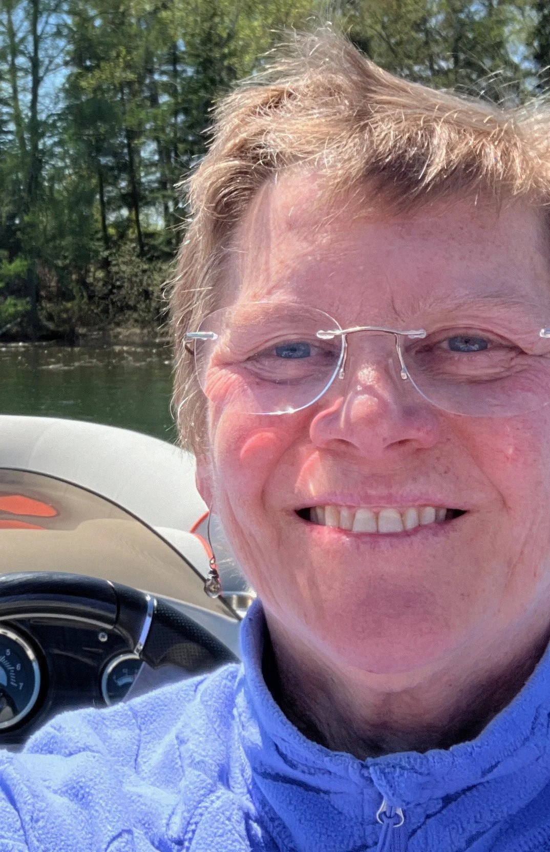 Close-up of a smiling person with glasses, outdoors on a boat, with water and trees in the background.
