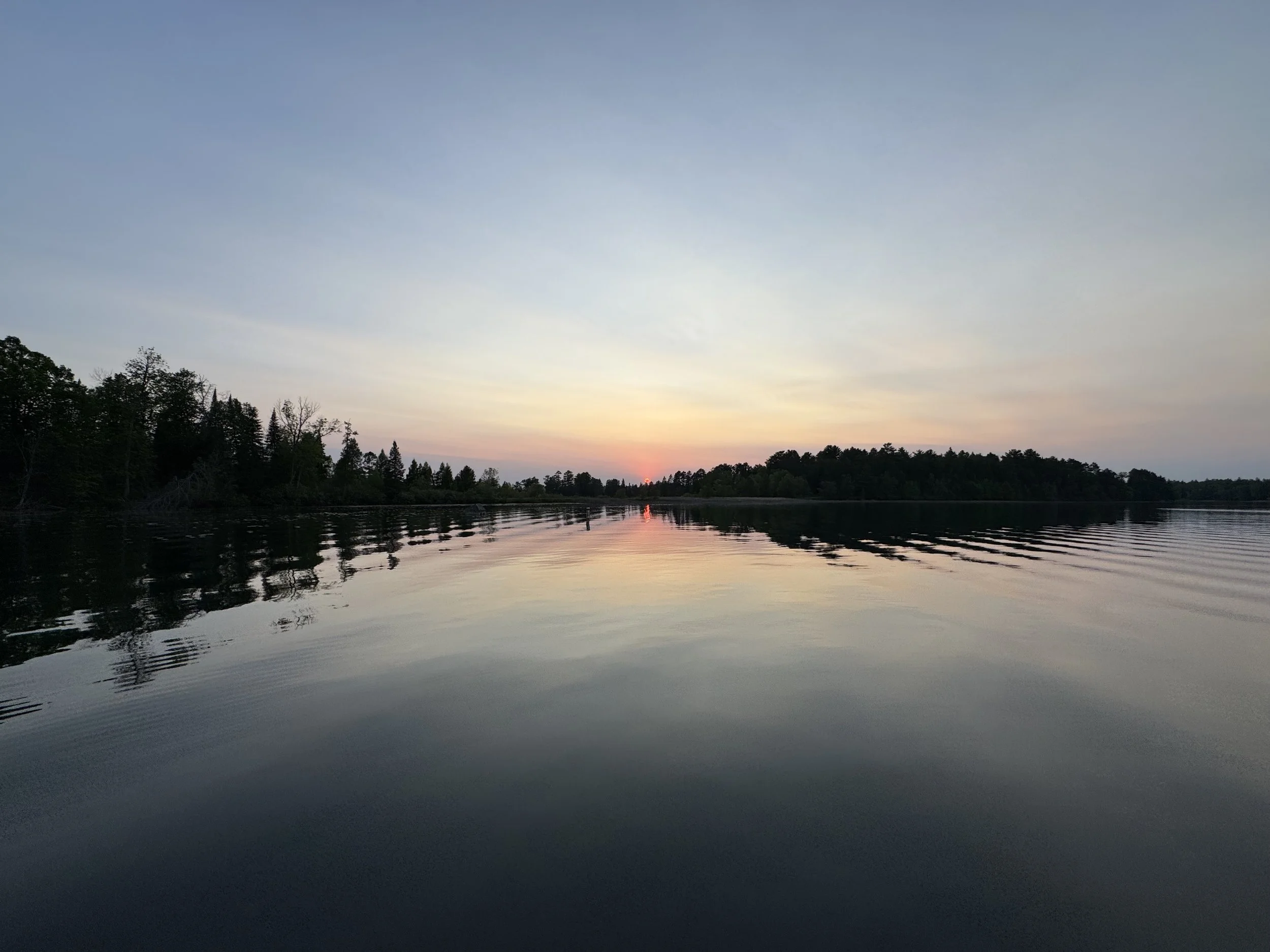 Calm lake at sunset with trees along the horizon and a reflection of the sky on the water.