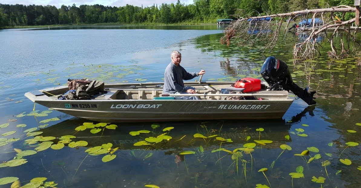 A man sitting in a small aluminum fishing boat on a lake with lily pads, near a fallen tree and boats in the distance.