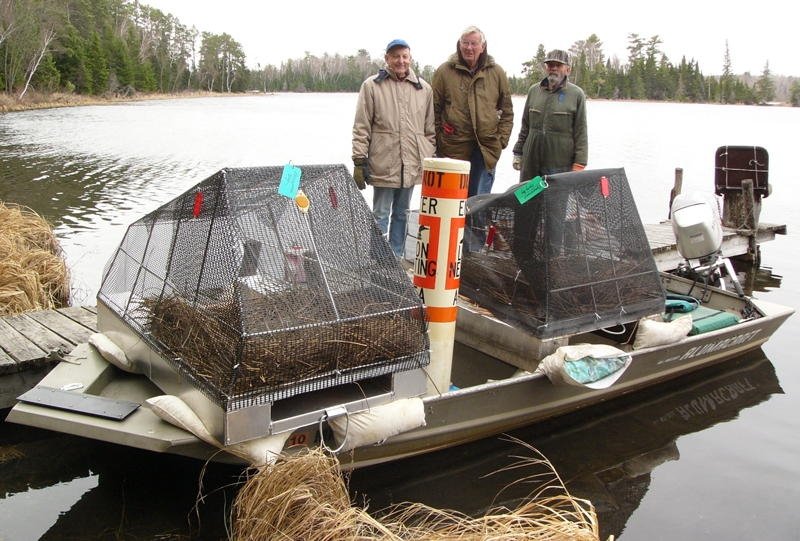 Three men standing on a dock beside a boat with two trap cages and a float underwater, surrounded by a forested lake.
