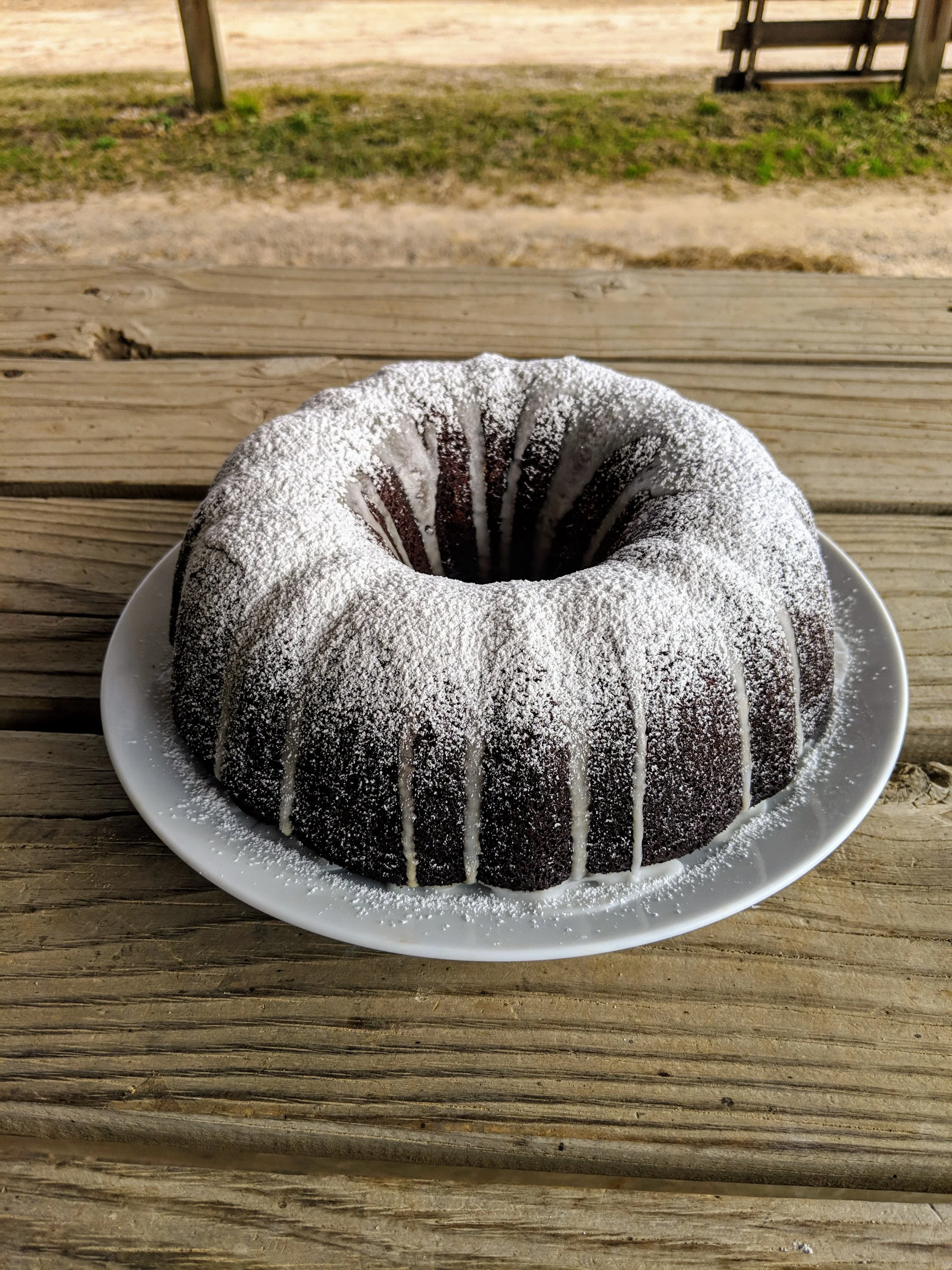 Chocolate bundt cake with white icing drizzle and powdered sugar on top, served on a white plate on a wooden outdoor table.