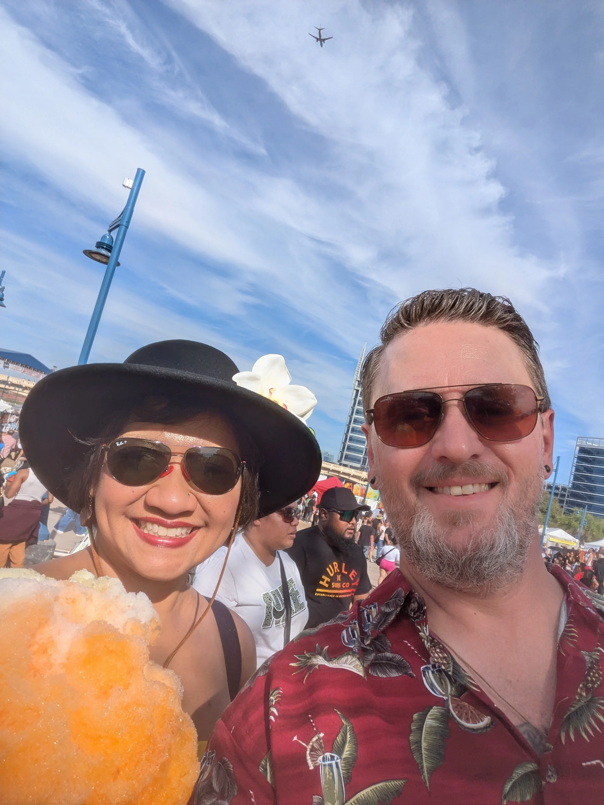 A smiling woman and man taking a selfie at a busy outdoor event. The woman is wearing a black wide-brimmed hat with a flower, sunglasses, and a white top, holding an orange-colored cotton candy. The man has a beard, short hair, sunglasses, and is wearing a red Hawaiian shirt with a floral pattern. The background shows other people, some with hats and sunglasses, a tall modern building, and a blue sky with some clouds. An airplane is visible flying overhead.