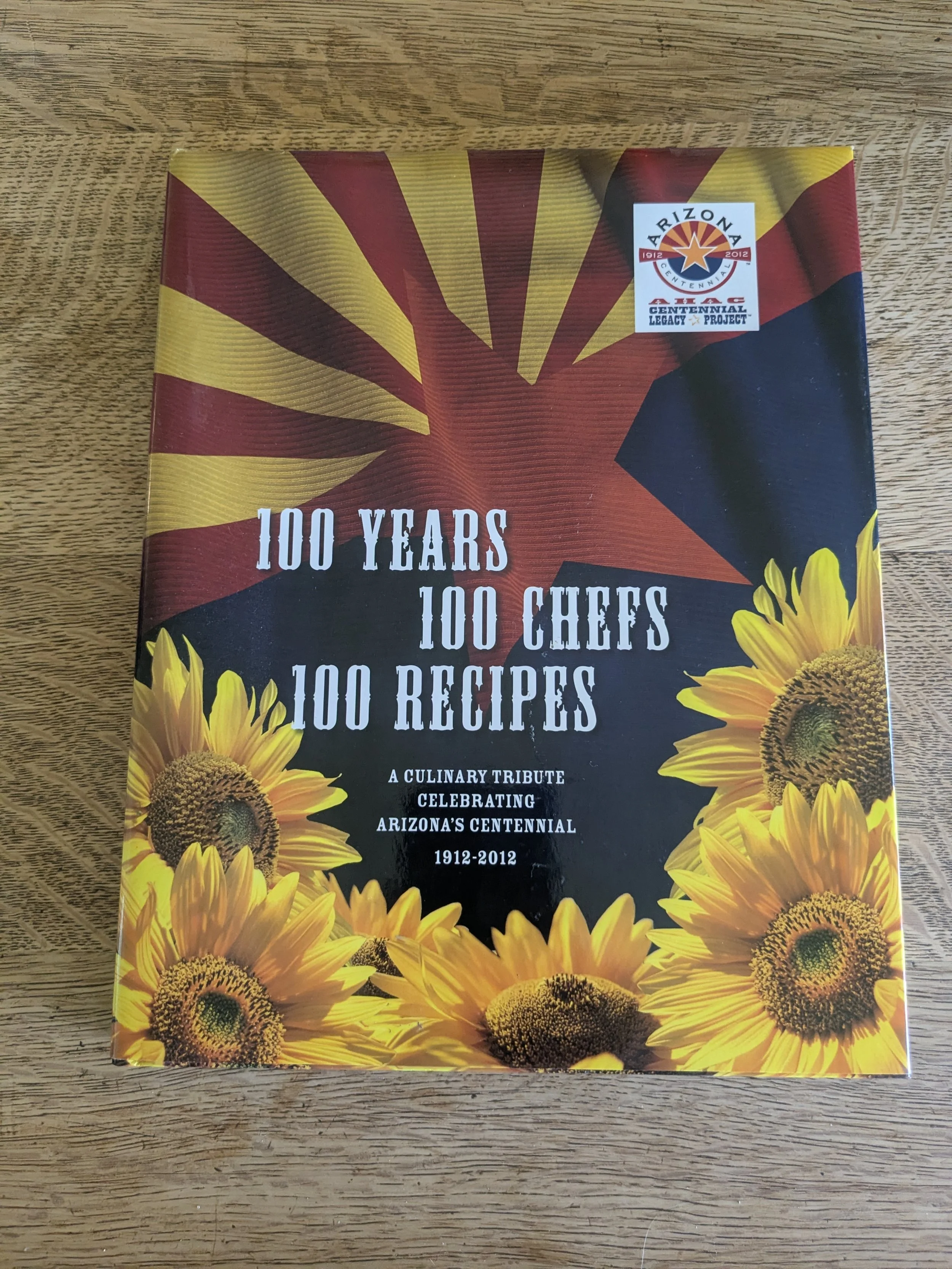 Book titled '100 Years 100 Chefs 100 Recipes' on a wooden surface, celebrating Arizona's centennial, with a sunflower design on the cover.