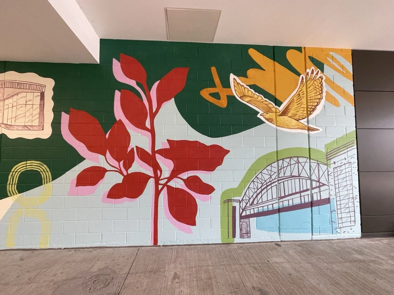 Section of completed mural on brick wall that featured bold, overlapping color blocks. In the middle of the mural, there is a large red plant with a pink outline. To the right of that, there is a big yellow-orange bird flying over a bridge.