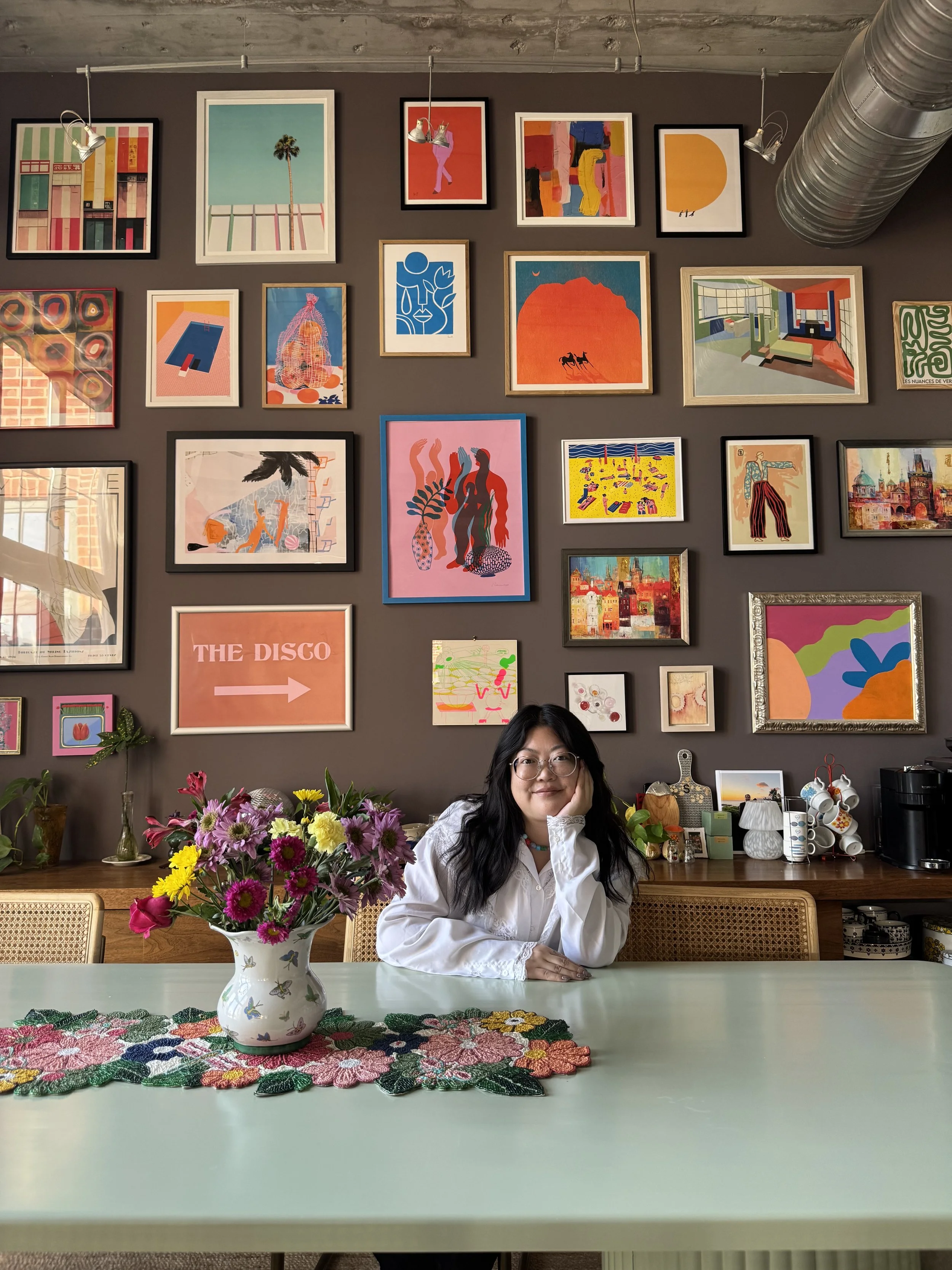 Sidney sits at her dining room table in front of her gallery wall.  Photo courtesy of Sidney Hui