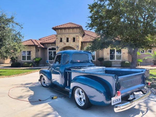 A vintage blue pickup truck parked on a driveway in front of a large house with a tile roof. The truck appears freshly painted with reflective shine, surrounded by green trees and well-maintained lawn.
