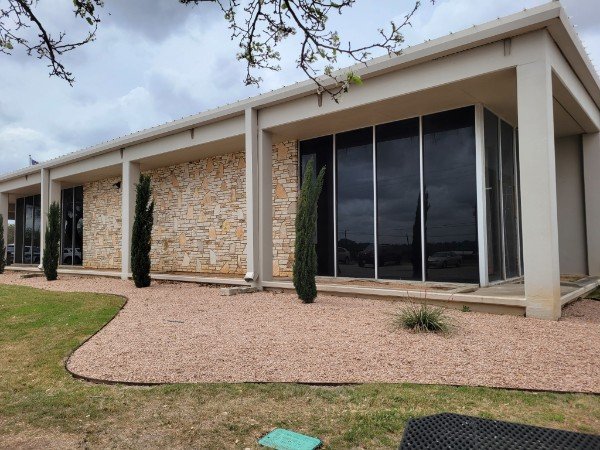 Modern single-story house with large glass windows, stone and white exterior walls, and a gravel yard with small trees and a grassy area.