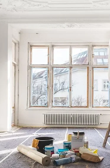 Room with large, bright windows, radiator beneath, and various painting supplies including paint cans and rollers on the floor.