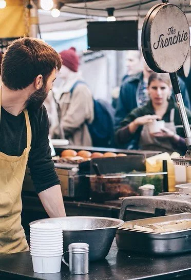 Person wearing an apron serving food at an outdoor market stall with "The Frenchie" sign, surrounded by cooking utensils and food items, while customers wait in line.