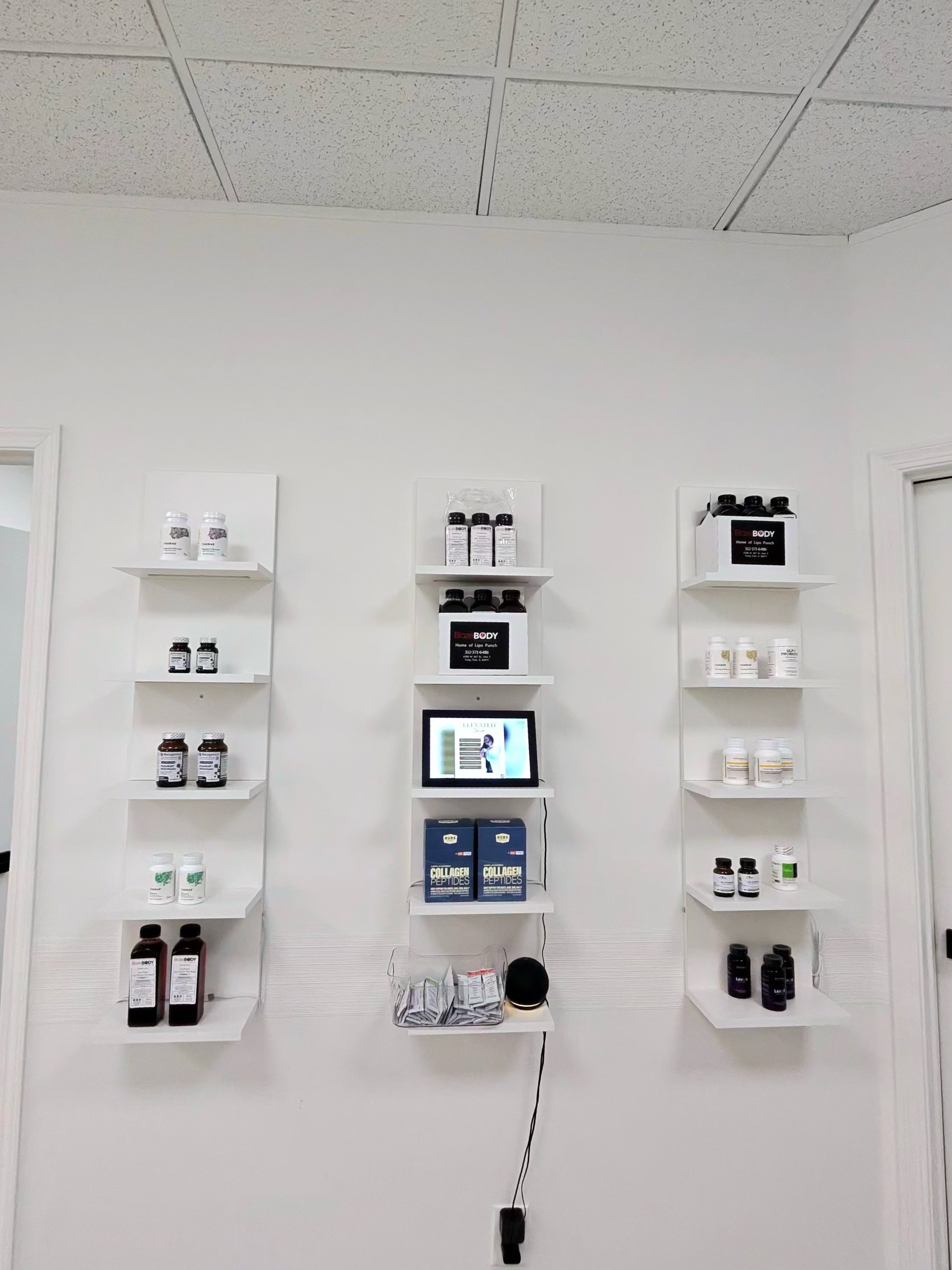 Retail display with shelves of supplement bottles, a computer monitor, and a basket of small items against a white wall and ceiling with tiles.