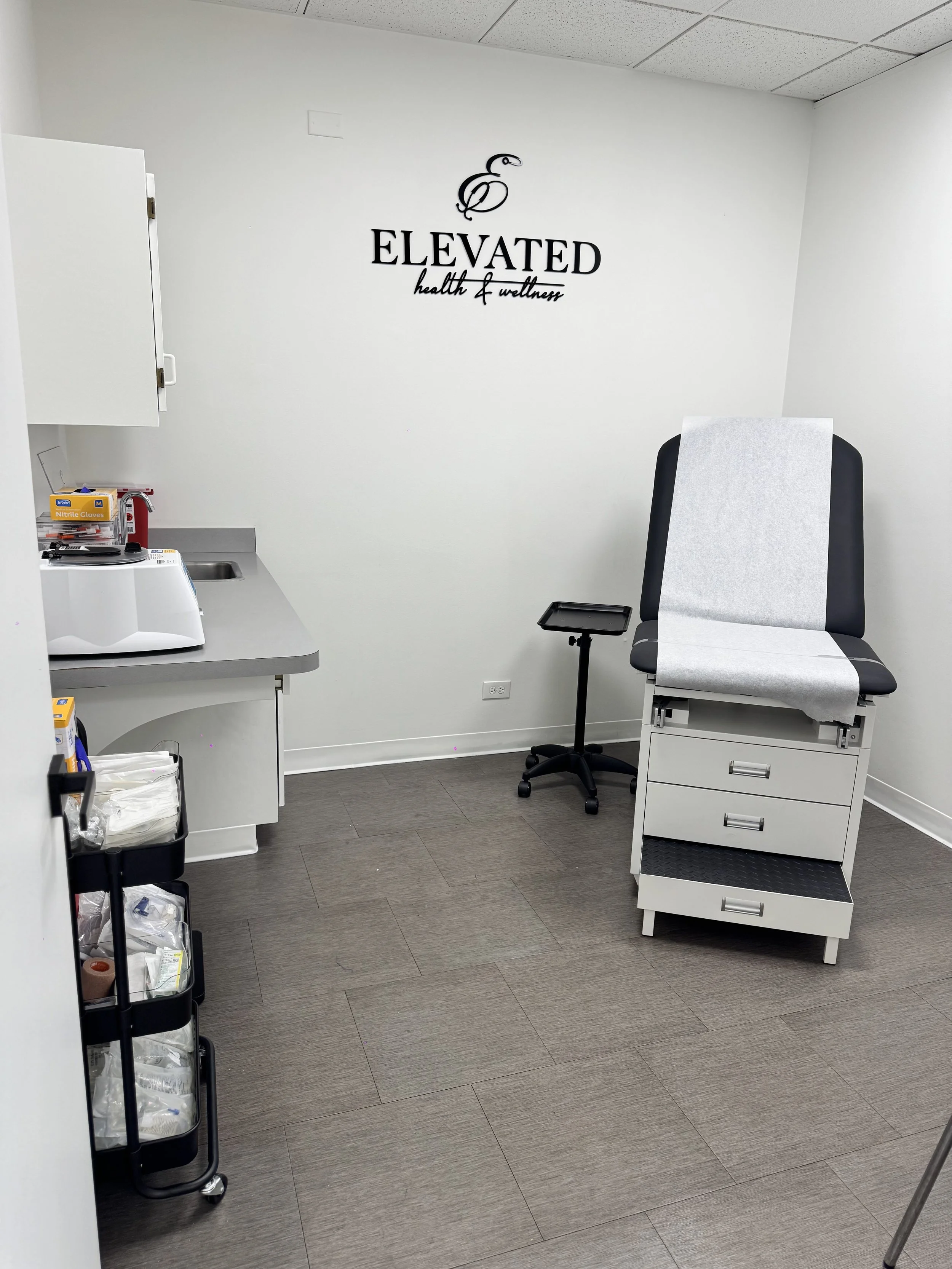 Medical examination room with a chair, a small table with a tray, and medical supplies on a cart. The back wall has a logo reading 'ELEVATED health & wellness' with a bird graphic.