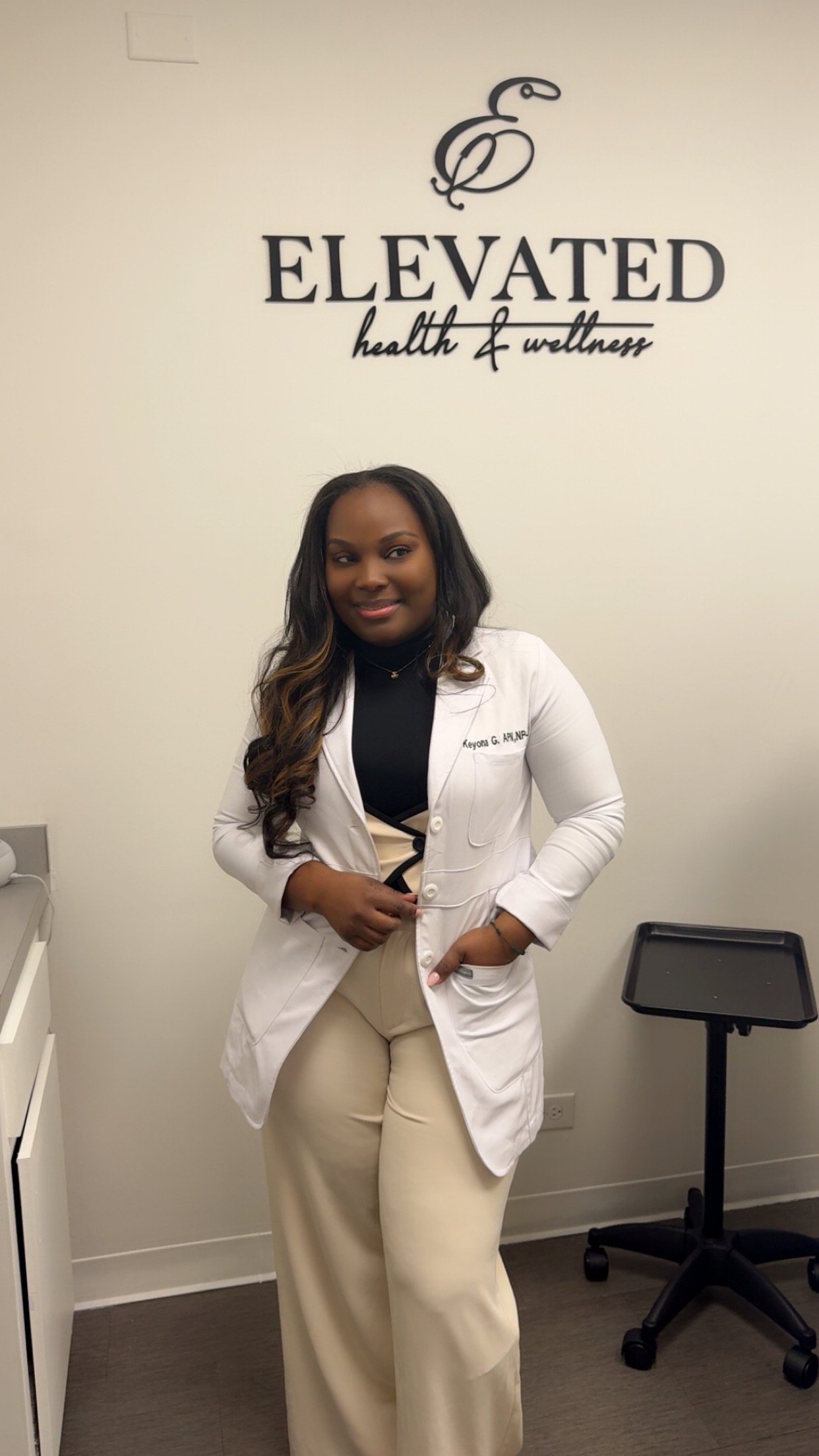 A woman standing in front of a wall with a logo that says "Elevated health & wellness," wearing a white coat, black turtleneck, and beige pants, smiling at the camera.
