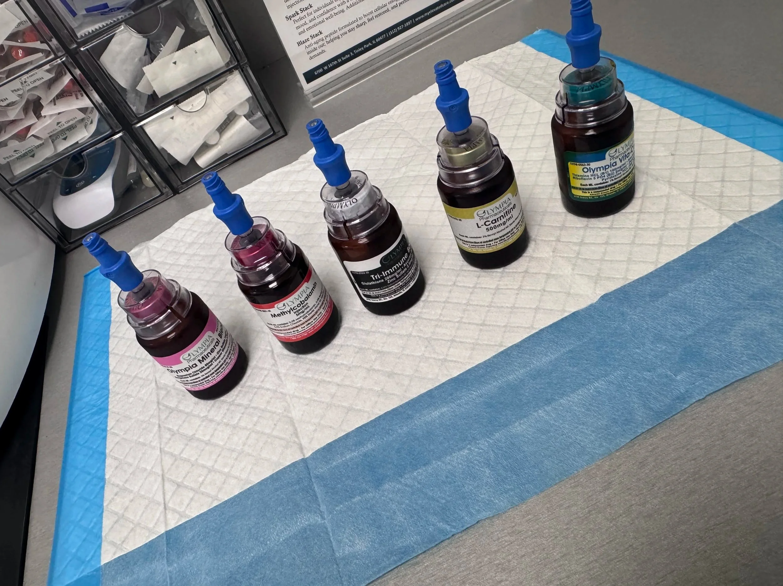 Four small bottles of medication with blue needle-like caps, placed on a paper pad on a gray surface, with a storage container in the background.