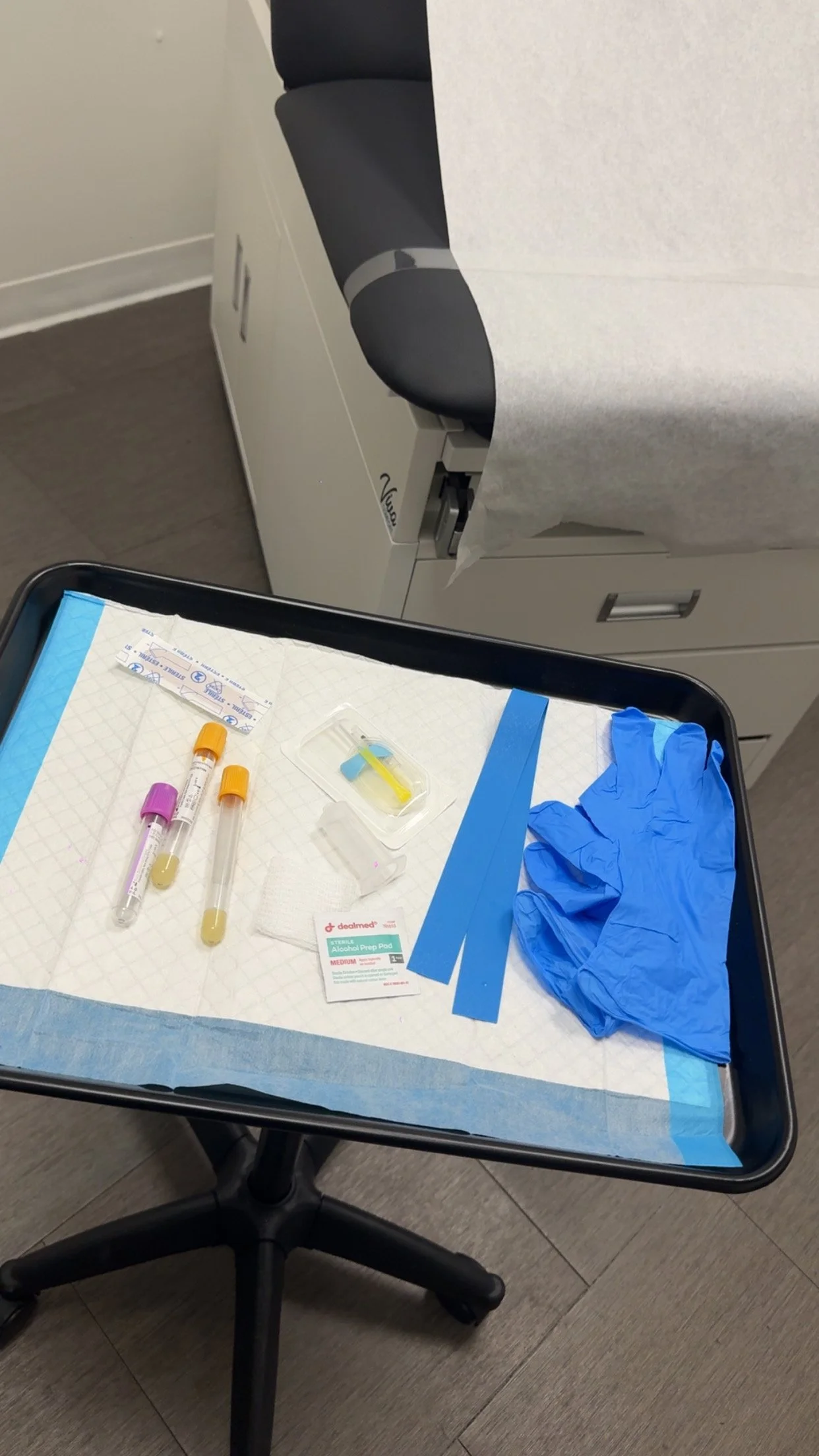 A tray with medical supplies including two test tubes, a pack of alcohol prep pads, a box of blue gloves, and various strips of blue tape. The tray is lined with a blue paper towel.
