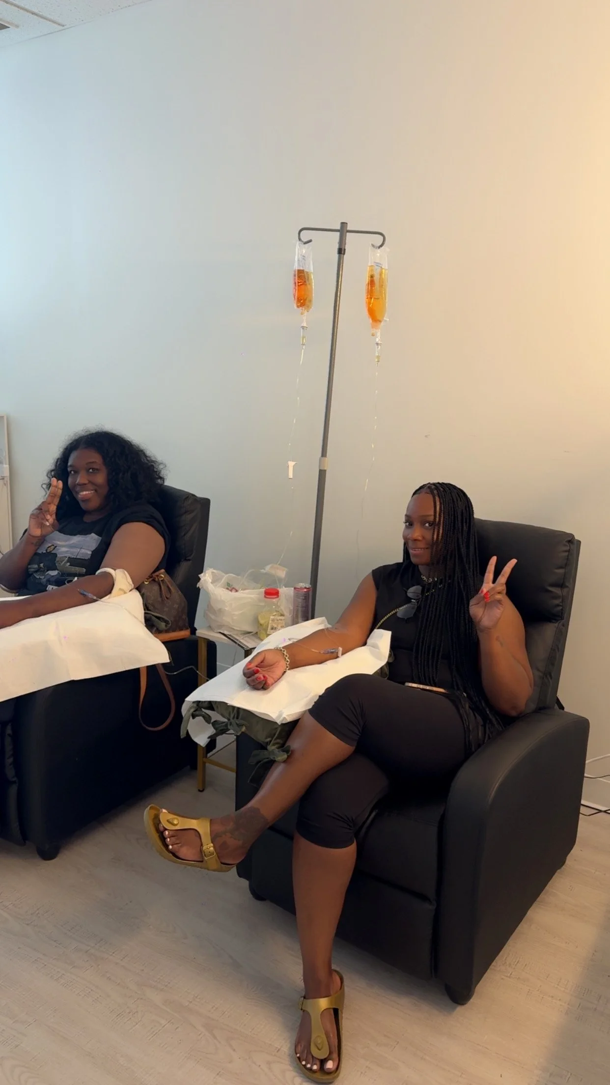Two women sitting in chairs donating blood, making peace signs with their hands, with intravenous tubing connected to their arms, and a stand with two IV bags hanging behind them.