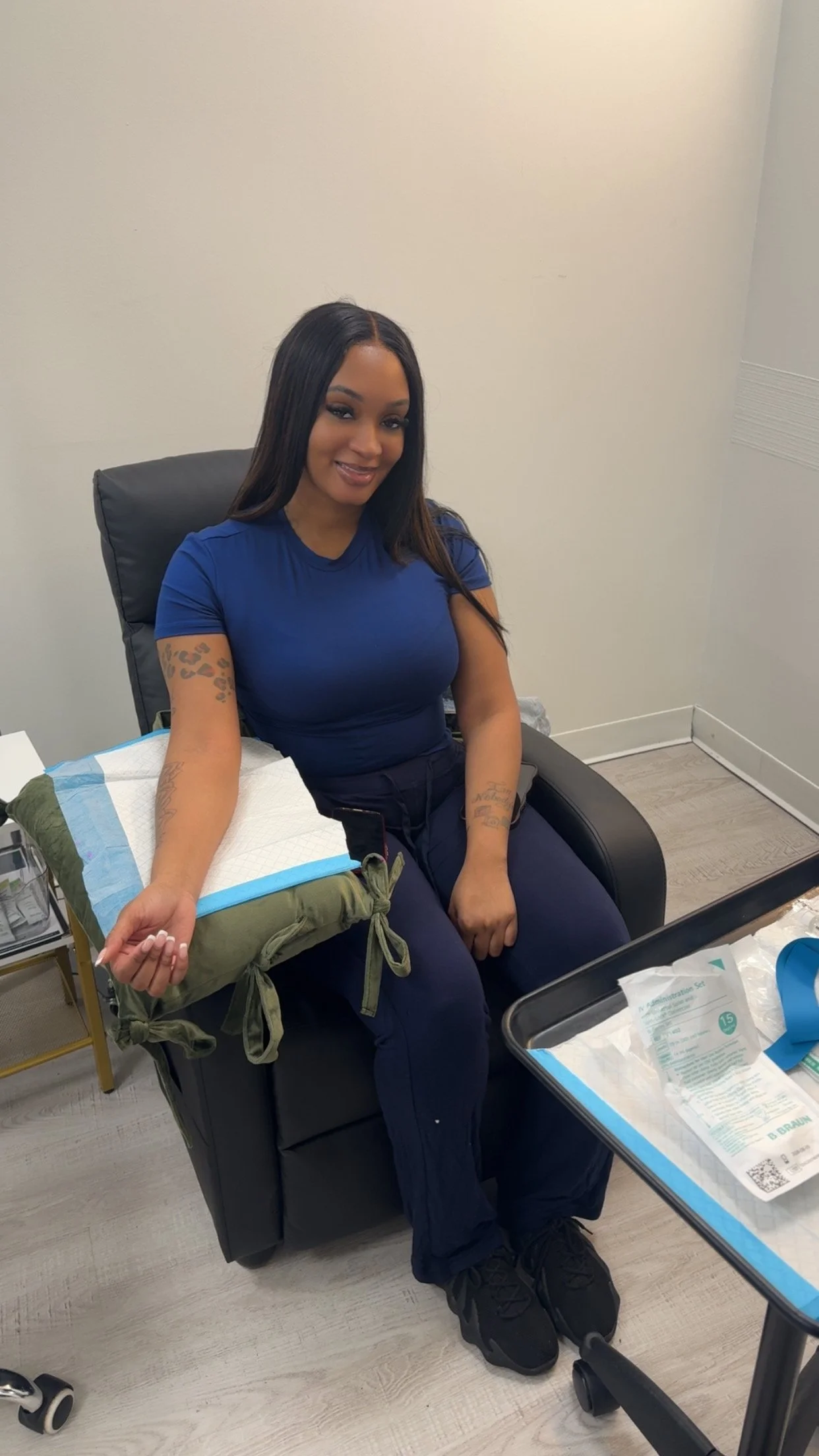 A woman sitting in a chair in a medical setting, preparing to donate blood. She has long dark hair, tattoos on her left arm, and is wearing a blue shirt and black pants. Medical supplies are on a tray nearby.