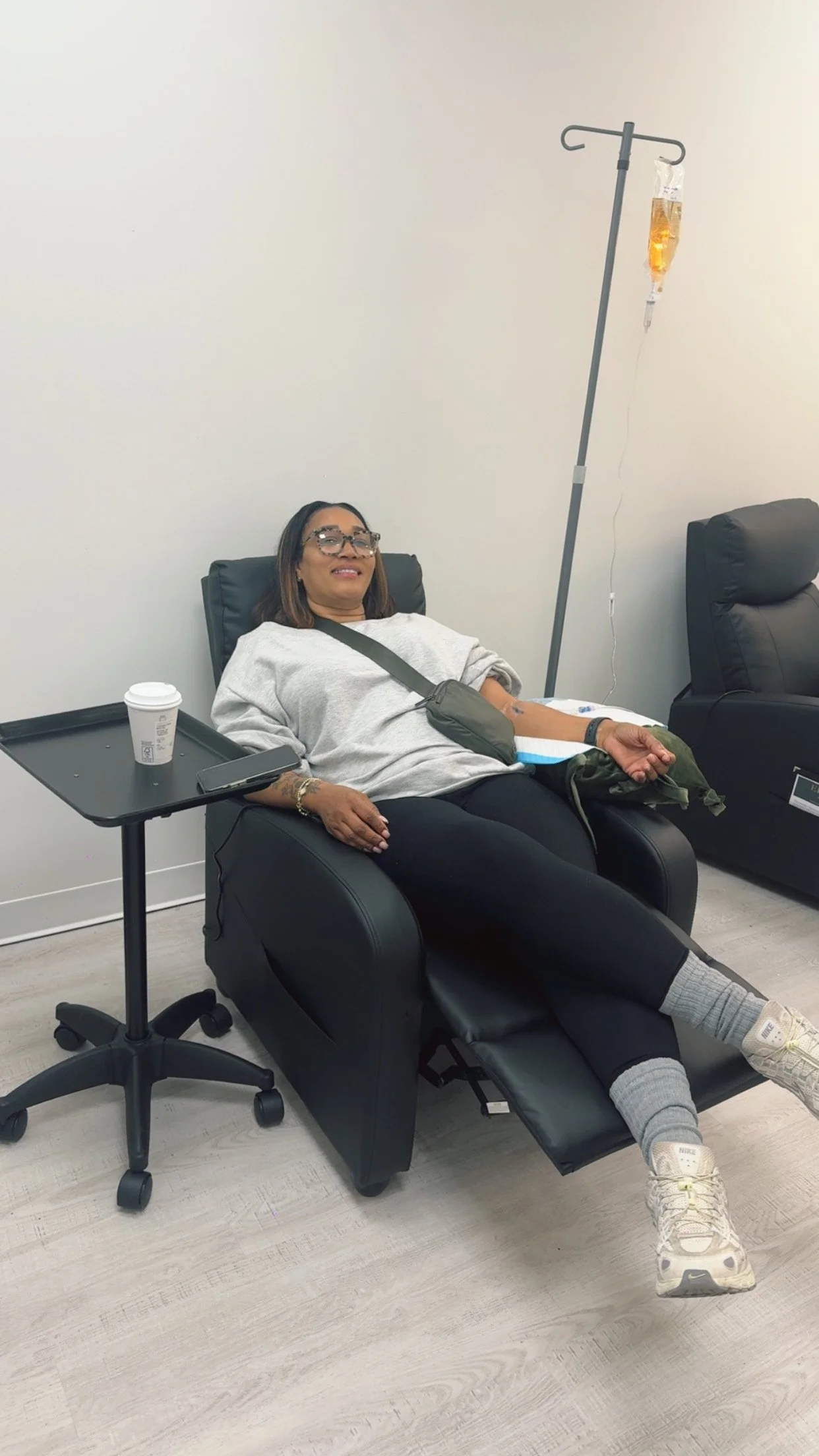 Woman donating blood in a chair at a medical facility, with an IV drip, a coffee cup, and a phone nearby.