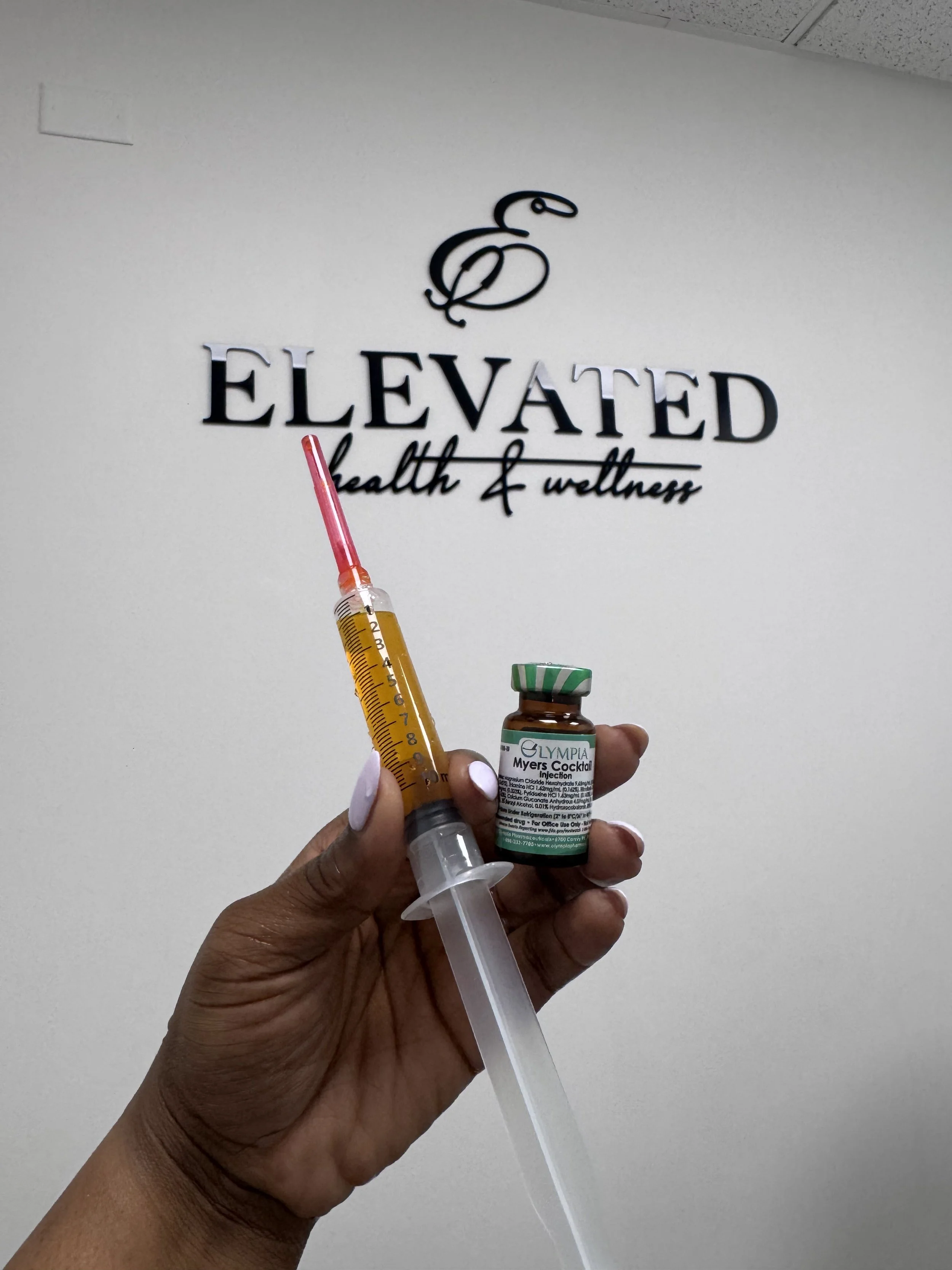 A person holding a syringe and a medicine vial labeled 'Myers Cocktail.' The background has a sign that reads 'ELEVATED health & wellness,' with a decorative letter 'E' above it.
