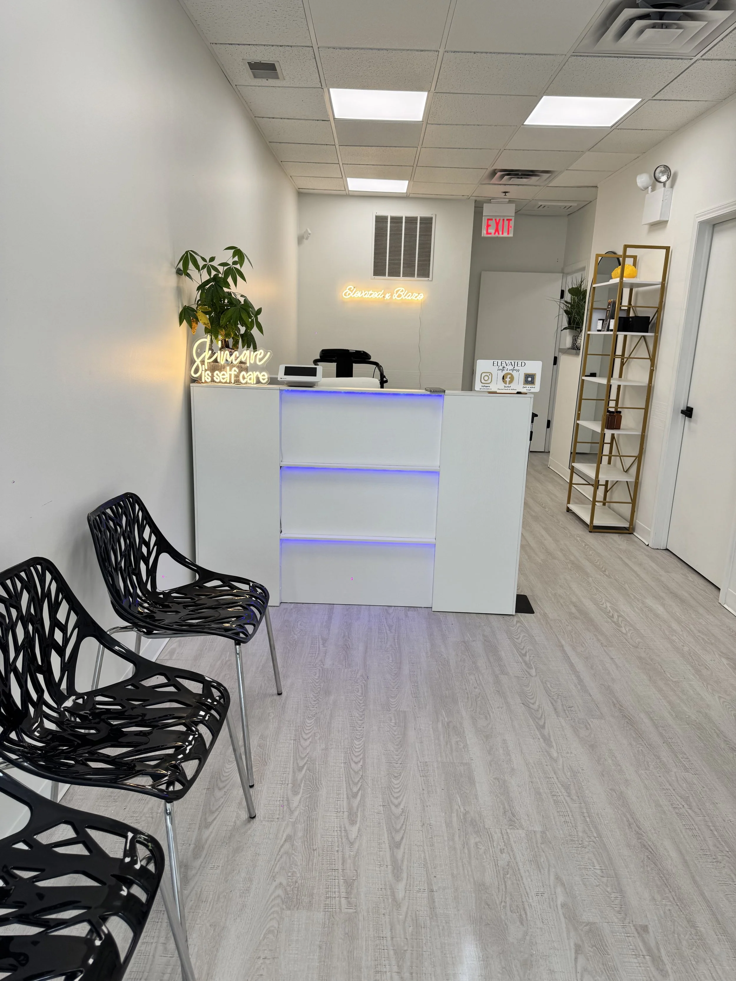 Reception area with black chairs, white counter with blue lighting, neon signs on wall reading 'Skincare is self care' and 'Elevated x Blaze', potted plants, and a gold shelving unit.