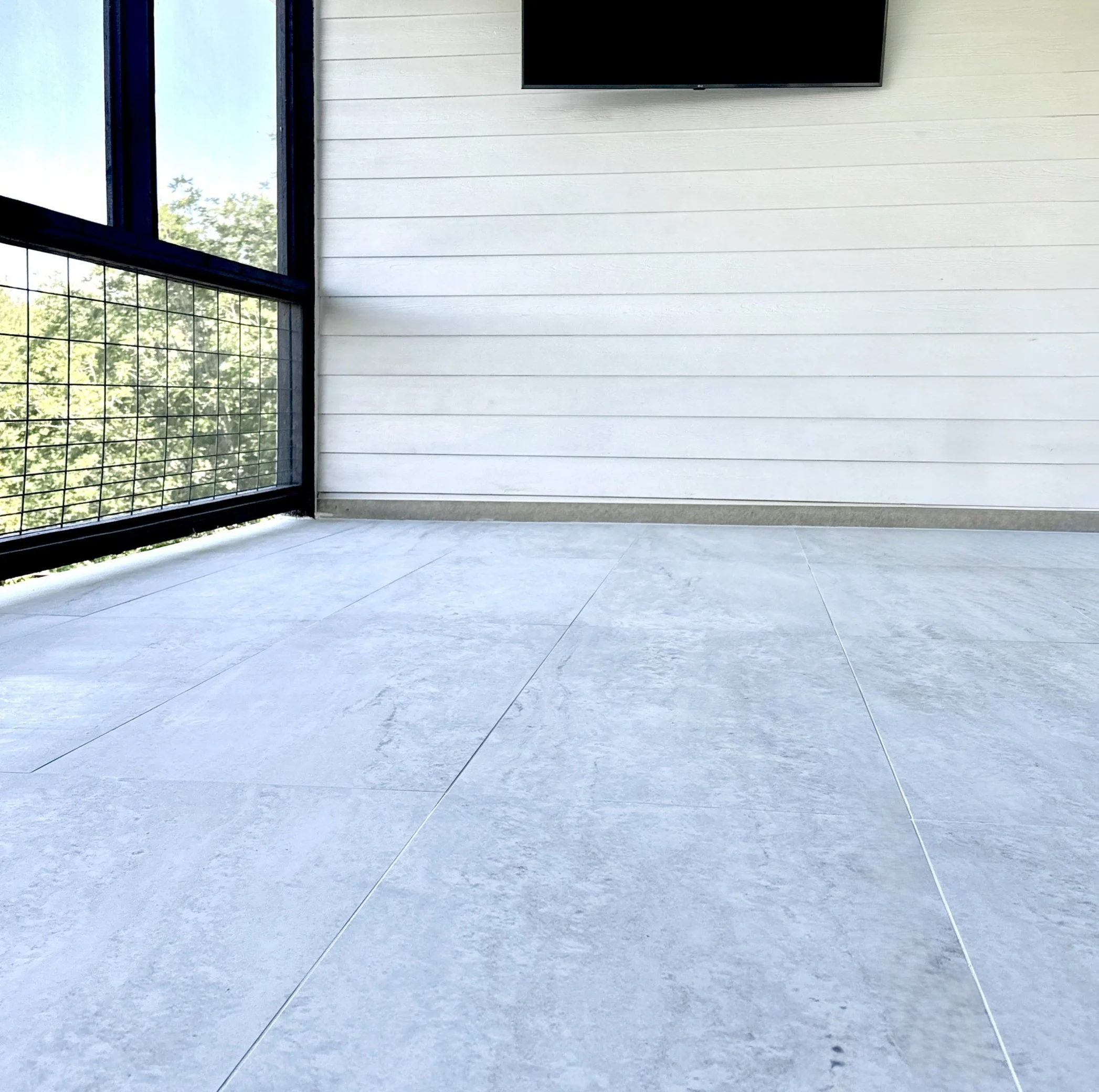 Exterior patio balcony with white tiled floor, white paneled wall, black window frame, and a small balcony railing with a view of trees outside.