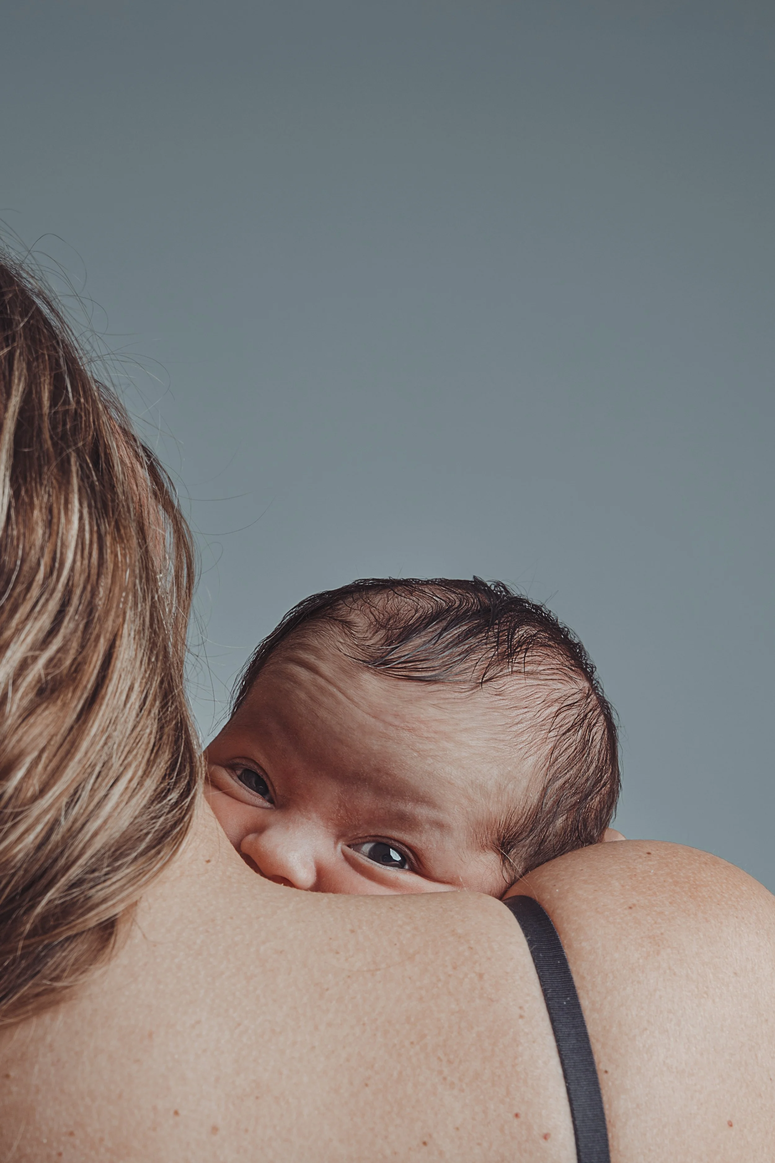 Close-up of a newborn baby resting on a woman's shoulder, looking at the camera, with a clear blue sky in the background.