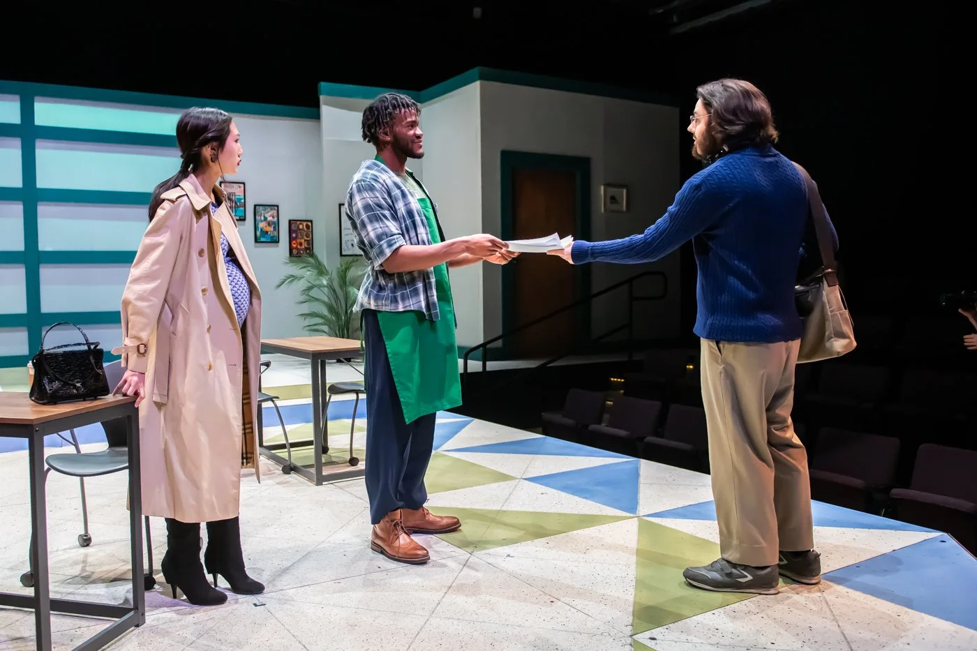 People on a stage with props for a play, featuring three individuals: a woman in a tan trench coat, a man in casual clothing with a green apron, and a man in a blue sweater, all engaged in a scene involving handing documents.