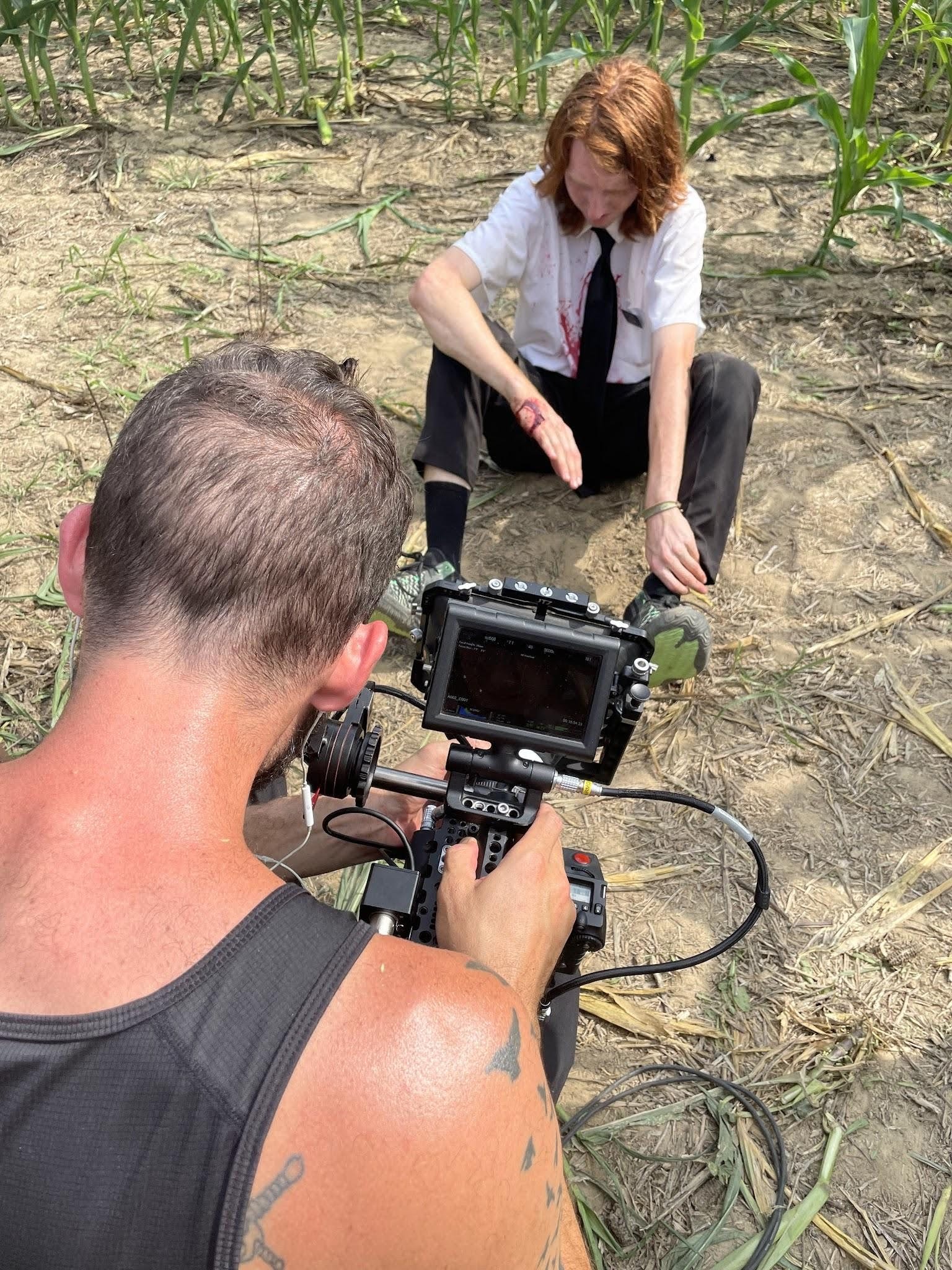 A person with tattoos is filming a woman sitting on the ground in a cornfield. The woman appears to be injured, with a bloodstained shirt and visible wounds on her arm, and she is in a stretching or reaching position.