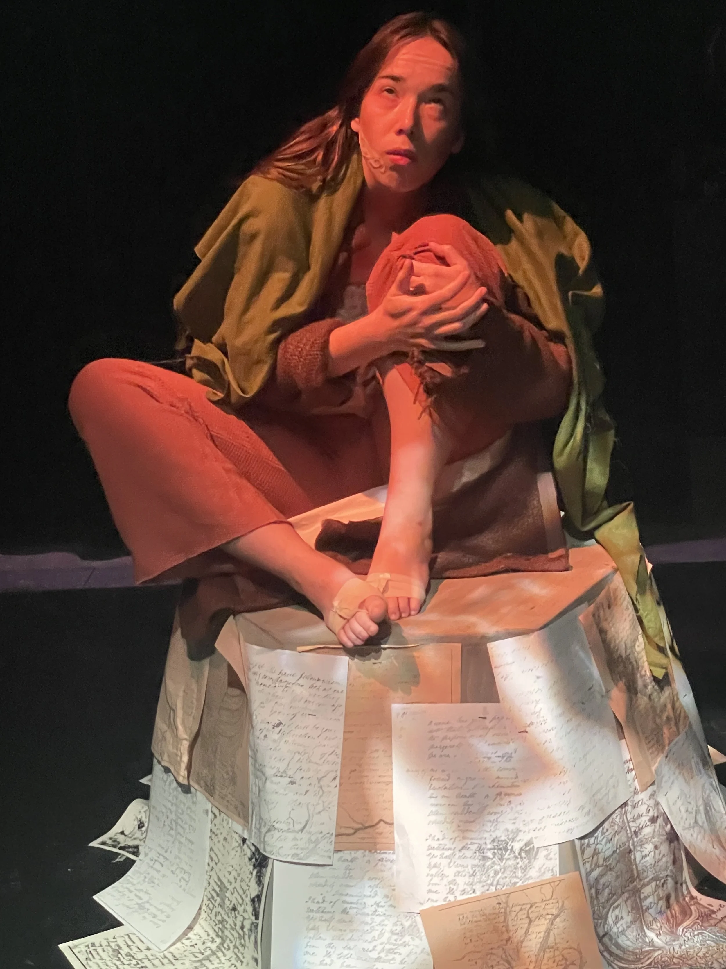 A theatrical scene with two women, one with long hair and one with a head covering, sitting on a table covered with handwritten notes and papers, in a dramatic pose.